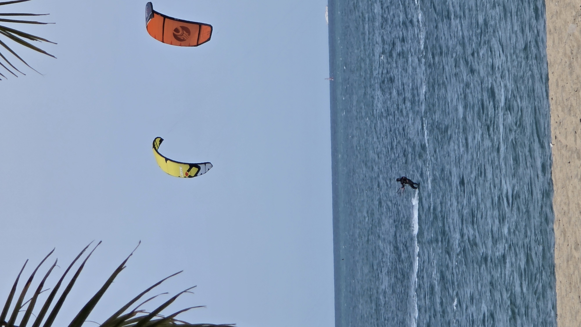 The image shows a beach scene with kite surfers in the water and kites in the sky. The location is likely Nha Trang Ward, Vietnam. A person is actively kite surfing, with the kite pulling them across the water. Two other kites are visible in the air, one orange and one yellow. Palm fronds are visible in the foreground, indicating a beach environment. The sea is a blue-grey color. A strip of sandy beach is on the right edge of the frame.