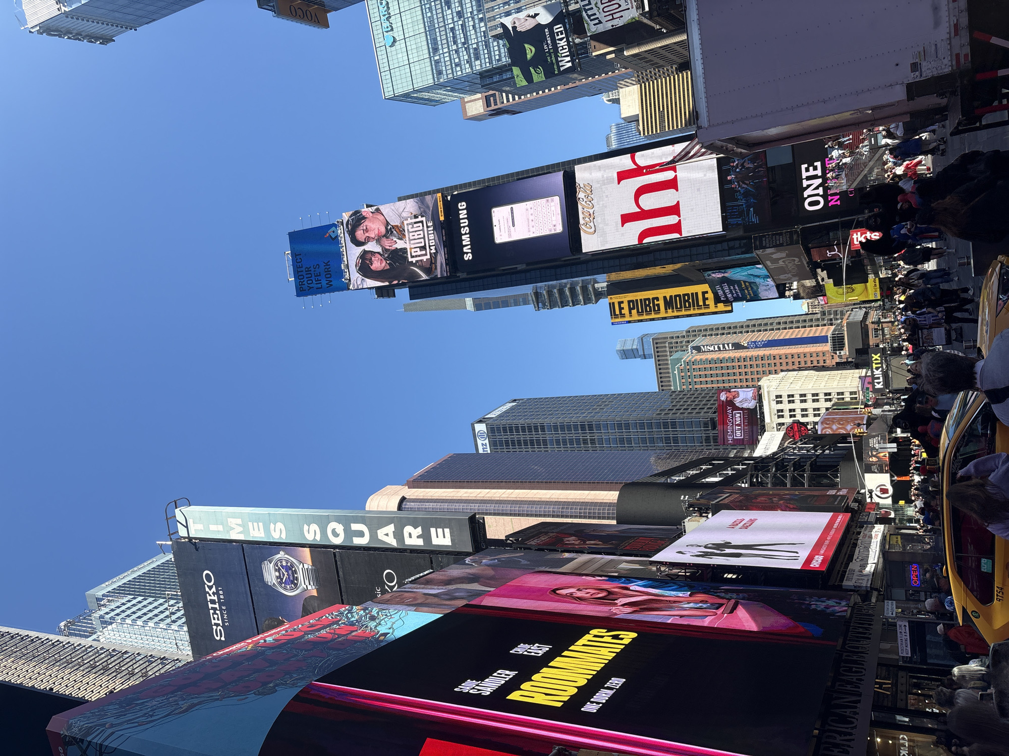 This image depicts Times Square in New York City, USA. The view is oriented upwards, showcasing the iconic billboards and skyscrapers. The billboards display advertisements for various brands and entertainment, including Samsung, PUBG Mobile, Coca-Cola, Wicked, and Roommates. People are seen walking on the sidewalks below, and a yellow taxi cab is visible. The sky is clear and blue, indicating a sunny day. The scene is characterized by the dense urban environment and the vibrant, commercial nature of Times Square.