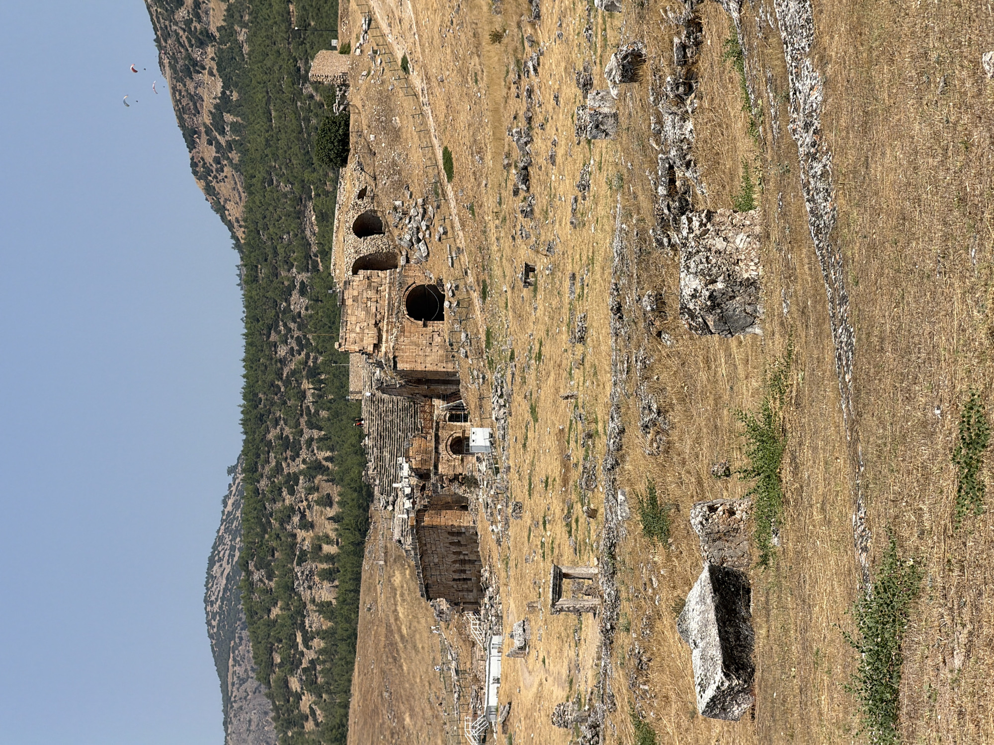 The image depicts a view of the ancient city of Hierapolis, located in Pamukkale, Turkey. The foreground is dominated by dry, golden-brown grasses and scattered ancient stone ruins, including large blocks and fragments. In the middle ground, the ruins of a large stone structure, likely a theater or amphitheater, are visible. This structure features arched openings and tiered seating arrangements, with some sections appearing partially collapsed. Several modern white structures and white railings with stairs are also present, indicating areas that have been preserved or are accessible to visitors. A dirt path winds through the ruins. In the background, a hillside covered with green trees rises, leading to a mountain range under a clear, pale blue sky. Several paragliders are visible in the sky above the mountains. The overall impression is of an archaeological site set within a natural, arid landscape.