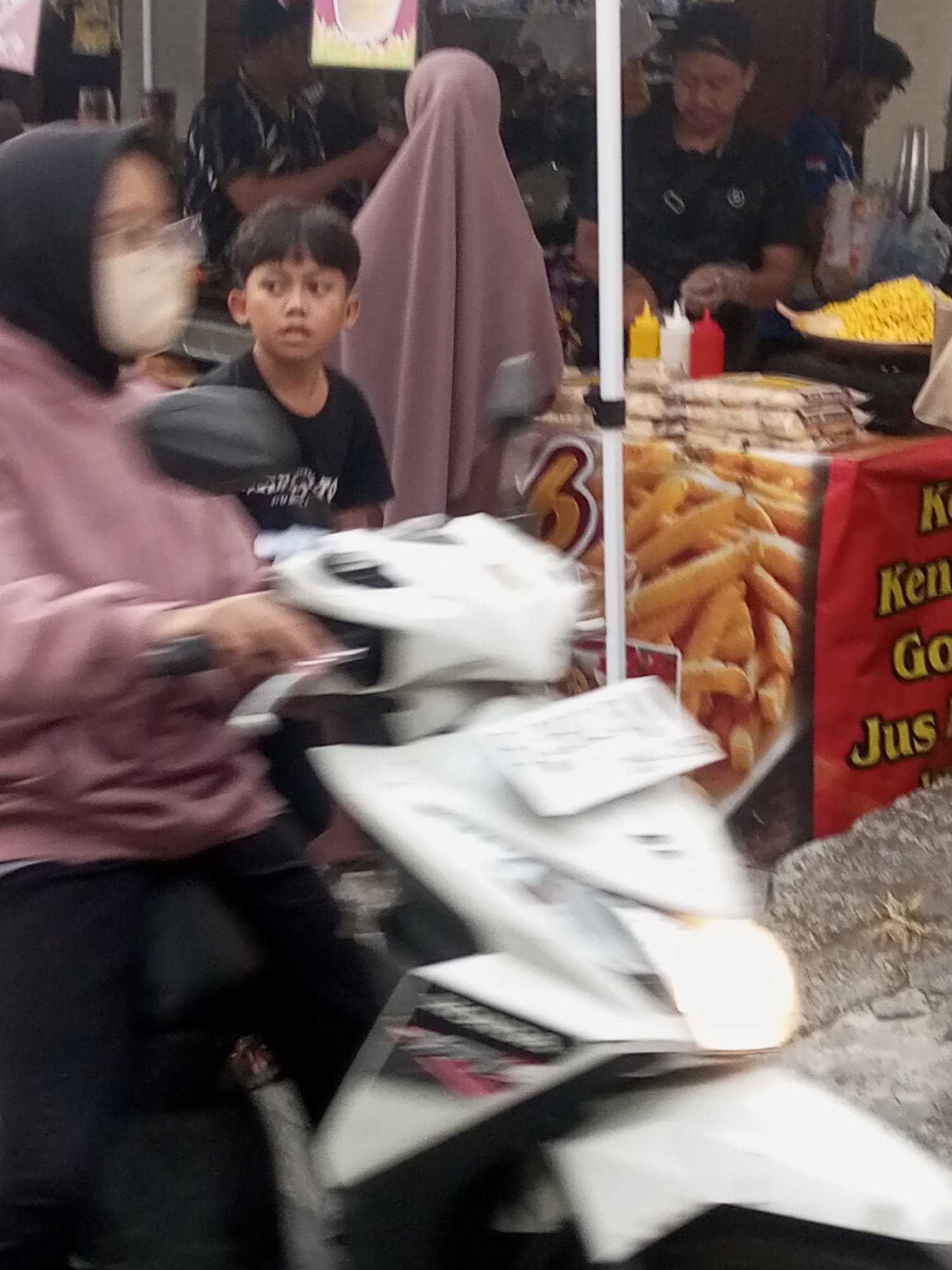 The image depicts a street scene in Pandansari, Indonesia, with a focus on a food stall. In the foreground, a person is seated on a white motorcycle, their upper body visible, wearing a pinkish hooded garment and black pants. To their right, a young boy in a black t-shirt looks to his left. Behind the boy, a person in a dark t-shirt is visible. The food stall features a display of french fries, with bottles of ketchup and mustard on the counter, and a yellow container holding what appears to be a snack. A red banner with Indonesian text, including "Ken" and "Jus," is visible on the right side of the stall. The environment suggests an outdoor setting, likely a market or roadside. The lighting is somewhat dim, with some artificial light sources contributing to the scene.