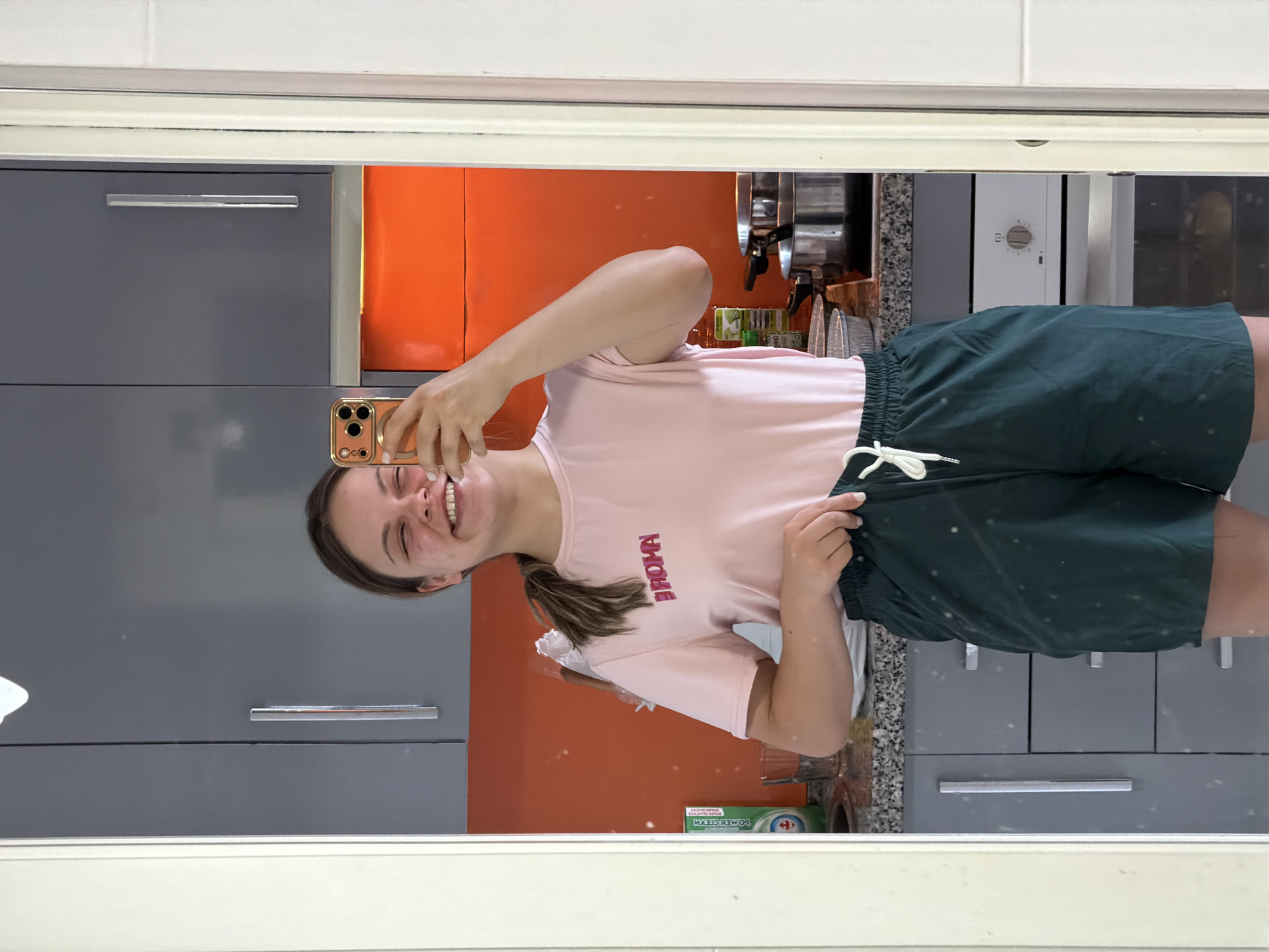 A young woman is taking a selfie in a bathroom mirror. She is wearing a pink t-shirt and green shorts. The bathroom has gray cabinets and an orange backsplash. A section of what appears to be a kitchen is visible in the background with stainless steel appliances. The location is Arroyo de la Miel-Benalmádena Costa, Spain.