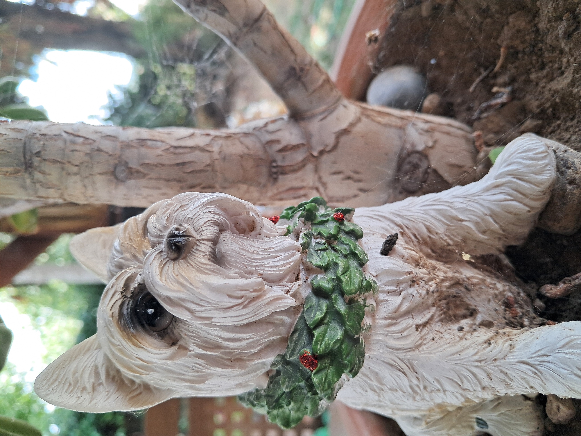A close-up, slightly high-angle shot features a ceramic statue of a white West Highland White Terrier dog wearing a green wreath with red berry accents around its neck. The statue is situated in a terracotta pot with dark soil and small stones. A thick tree trunk with rough bark stands to the right of the statue, partially obscuring the background which consists of blurred green foliage and a bright, overexposed sky. Spiderwebs are visible across the scene, particularly between the statue and the tree trunk. A small, dark insect is resting on the statue's chest. The lighting suggests an outdoor setting, likely during the daytime. The statue appears aged and somewhat dirty, with brown staining on its lower body and paws. The overall scene is static and depicts a still-life arrangement.