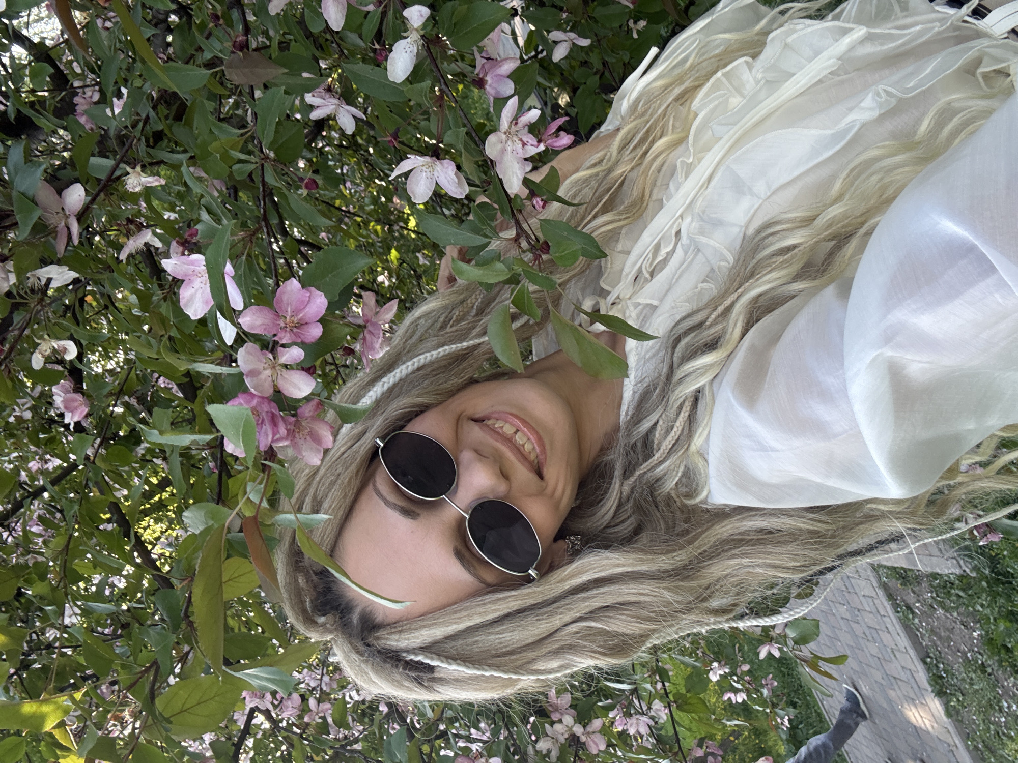 A woman with long, blonde hair styled in braids and wearing round sunglasses is lying down amongst the flowering branches of a tree. She is smiling and wearing a white, layered top. The tree has pink and white blossoms and green leaves. In the background, there is a glimpse of a brick pathway and green grass. This image might be taken in Rusanovo, Russia, based on the request.