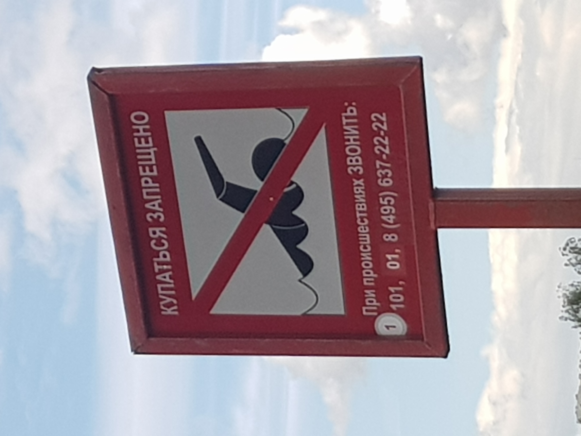 A red sign with white text in Russian reads "КУПАТЬСЯ ЗАПРЕЩЕНО" (Swimming is prohibited) and "При происшествиях ЗВОНИТЬ:" (In case of emergency, call:) followed by the phone numbers 101, 01, and 8 (495) 637-22-22. The sign displays a black figure of a swimmer with a red diagonal line across it, indicating that swimming is not allowed. The sign is mounted on a pole, and the background shows a partly cloudy sky. The location is Unknown, Unknown.