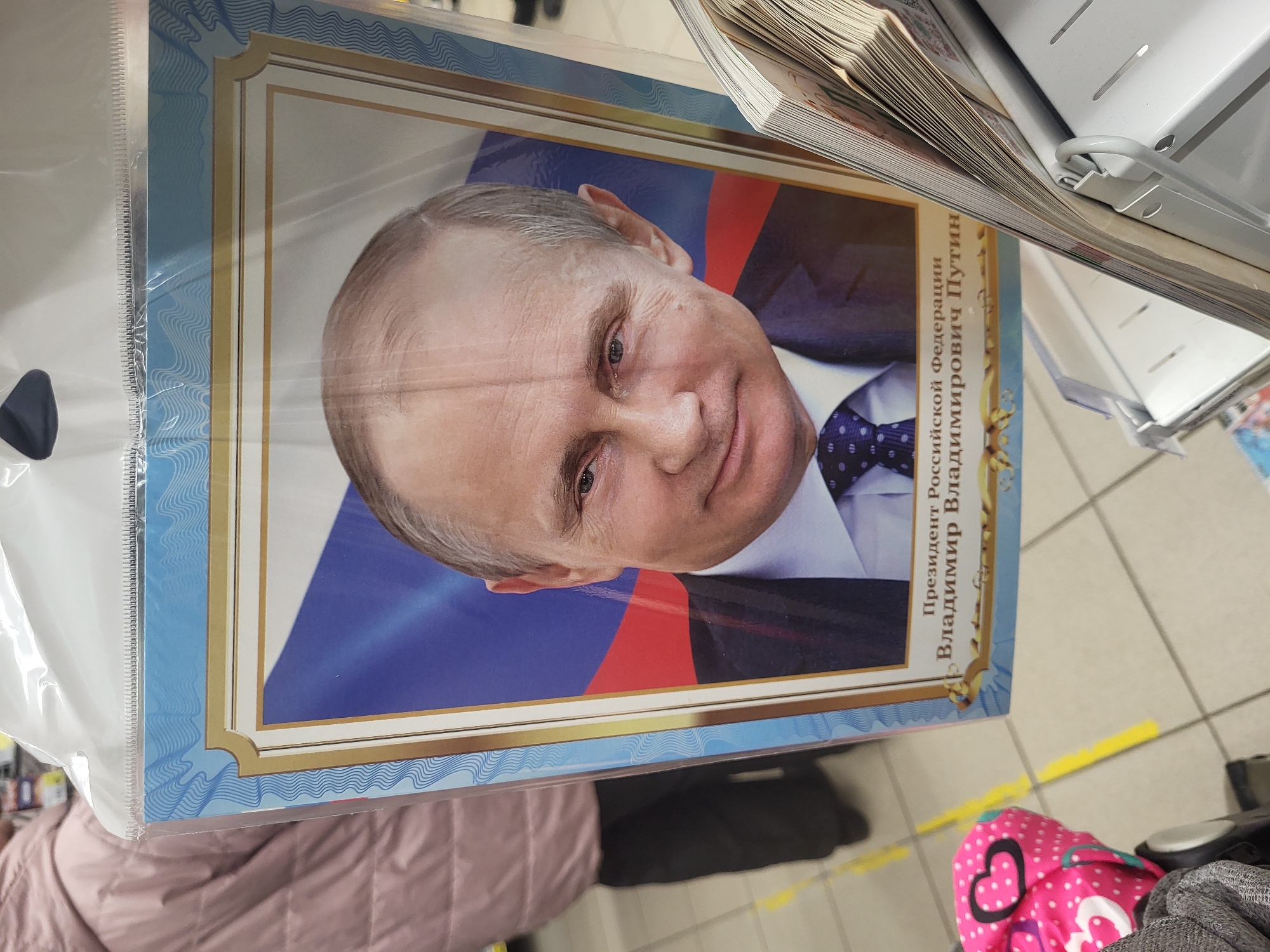 A medium-close shot shows a framed picture of Vladimir Putin on a shelf in a store. The picture is in a clear plastic wrapping and features Putin's face against the backdrop of the Russian flag, with the text "Президент Российской Федерации Владимир Владимирович Путин" (President of the Russian Federation Vladimir Vladimirovich Putin) printed next to his image. The frame has a light blue border with decorative gold lines. The surrounding context includes stacks of papers or magazines above the picture. The lower part of the picture and the frame are the subject of the shot. In the foreground and background, portions of clothing and footwear suggest a shopping environment, as well as a tiled floor with a yellow stripe on the tile. The photo was taken in the city of Chaikovsky, Russia.