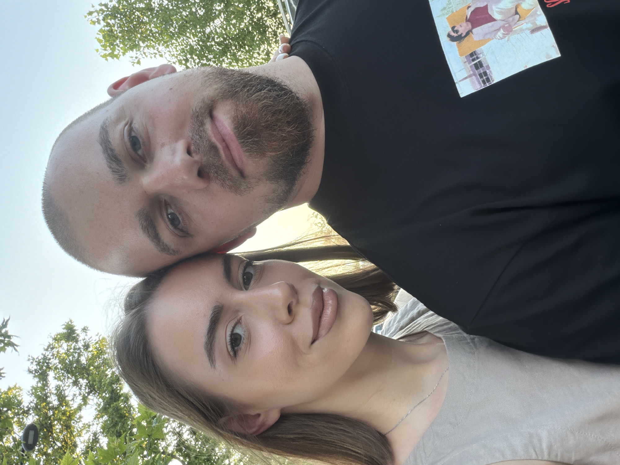 This is a selfie taken outdoors, likely during the daytime under a clear sky, given the bright lighting. The image features two people: a man and a woman, positioned close together. The man's head is tilted down towards the woman, with his forehead gently touching hers. He has a short haircut and a beard, and appears to be wearing a black t-shirt with a graphic print on it. The woman is looking towards the camera, with her head angled slightly. She has long brown hair and appears to be wearing a light-colored top with a delicate silver necklace. The background includes foliage, suggesting a natural or park-like setting. The overall tone of the image is intimate and happy, capturing a moment between two people. A printed image is visible on the man's shirt, but the details within it are not clearly discernible. There is no other visible text in the image. Based on the provided location context, the scene is set in Nesebar, Bulgaria.