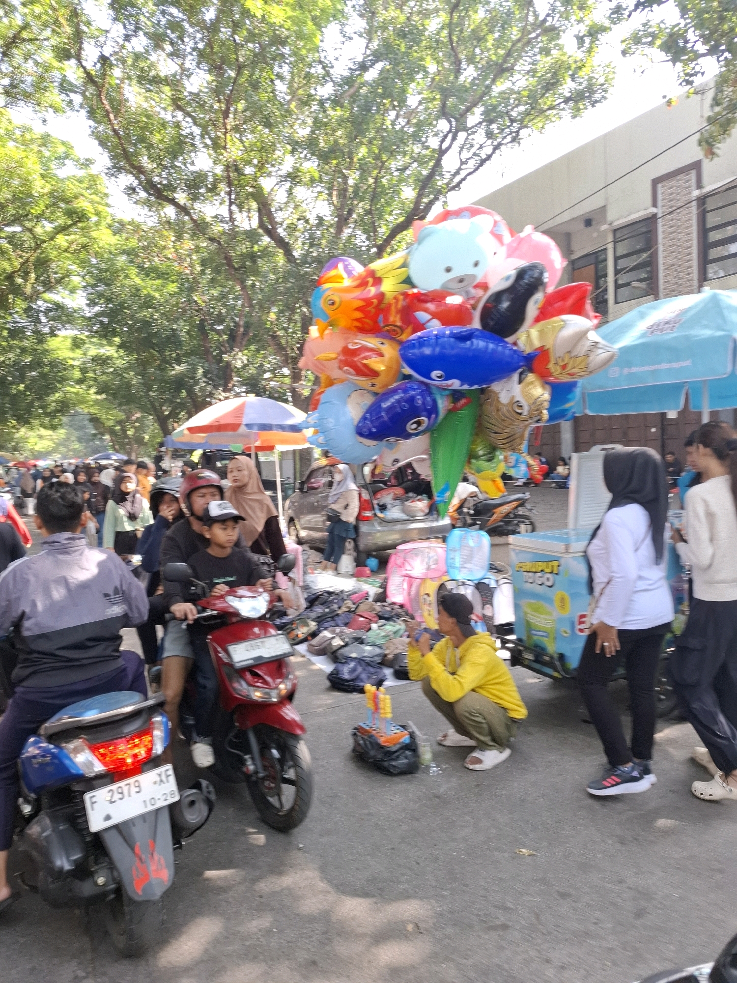 In Karangtengah, Indonesia, a street scene unfolds with a vendor selling a large cluster of colorful balloons, predominantly featuring animal shapes like fish and a bear. The balloons are tethered and rise above a busy street market. In the foreground, two motorcycles are parked, one blue and one red, with riders. A person in a yellow jacket is crouching near the balloons, possibly a buyer or assistant. Several other individuals are walking or standing around the market stalls, which are partially visible and display various goods including clothing and bags. A blue and white ice cream cart is also present. The environment appears to be outdoors with trees and buildings lining the street, suggesting a commercial area on a sunny day.