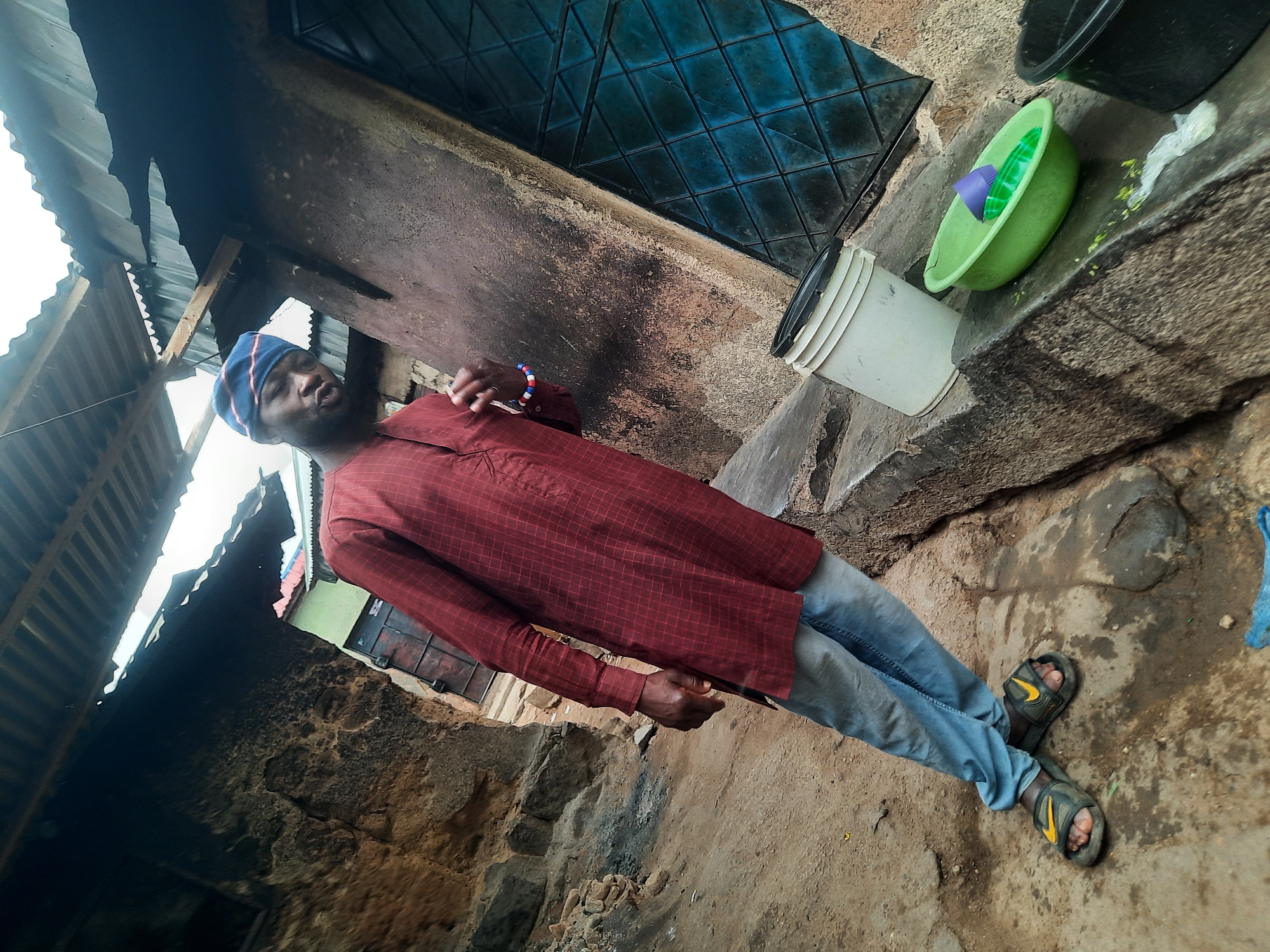 A man stands outdoors in Gwong, Nigeria. He wears a red, long-sleeved, plaid shirt, blue jeans, and dark-colored sandals with yellow logos. He has a blue and white head covering. The setting appears to be a narrow alleyway or walkway between buildings. A corrugated metal roof overhangs on the left. An exterior wall is on the right with a blue window, a white bucket, and a green bowl containing purple and green objects. The ground is dirt.
