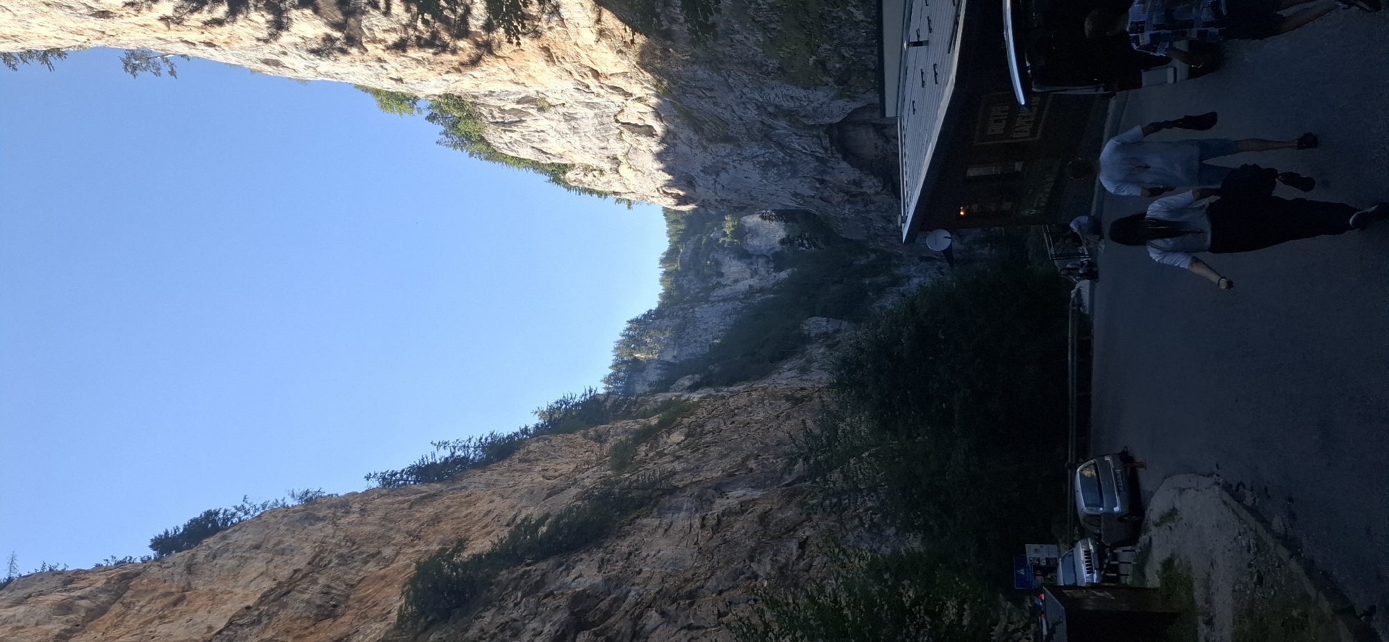 The image depicts a dramatic gorge with steep, rocky cliffs on either side, partially covered in vegetation. The sky is clear and blue. In the foreground, a paved road curves through the gorge. On the right side of the road, a building with a dark roof and wooden walls is visible, displaying Cyrillic text that appears to be an advertisement for a restaurant. Several cars are parked along the left side of the road. Four individuals are walking on the road, heading away from the camera and into the gorge. The lighting suggests it is either early morning or late afternoon. This scene is located in the Trigrad Gorge, near the village of Trigrad, Bulgaria, a region known for its natural beauty and karst formations.