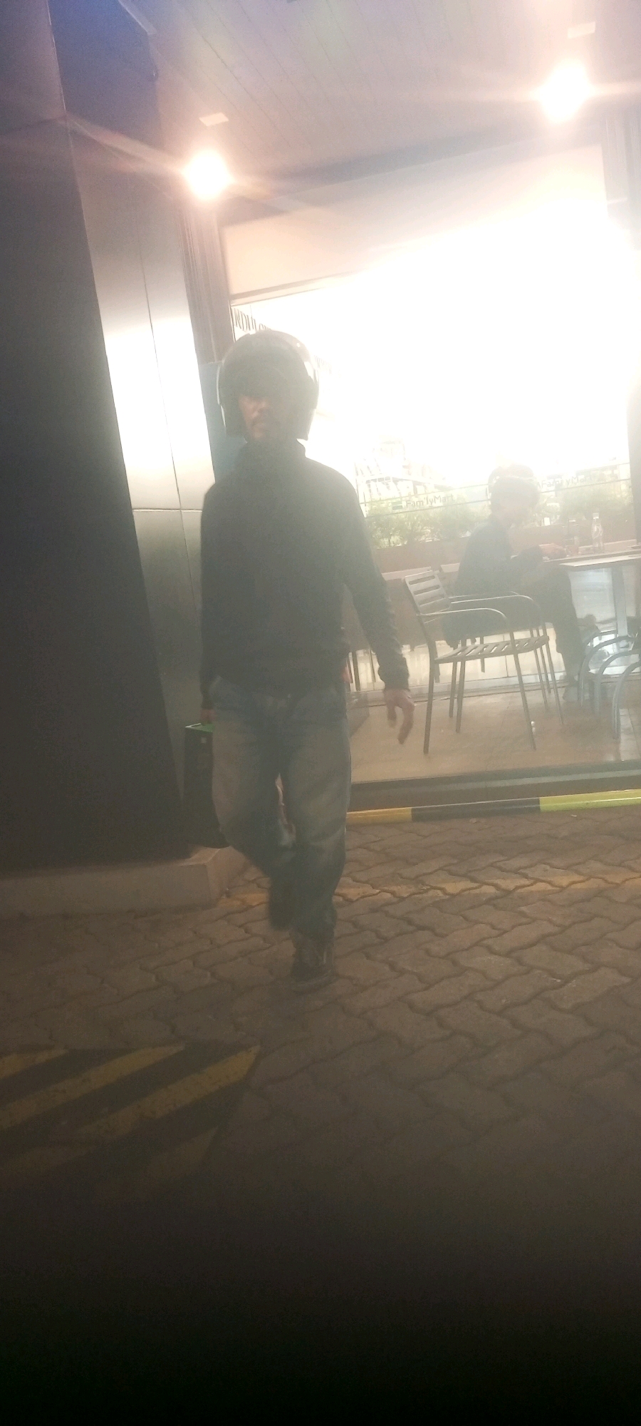 A person wearing a black motorcycle helmet and a dark long-sleeved shirt walks across a paved outdoor area. The person is wearing blue jeans and dark shoes. The pavement is made of interlocking bricks and has yellow and black striped lines on it. In the background, there is a glass-enclosed area with tables and chairs, suggesting an outdoor seating area for a establishment. Another person is visible sitting at one of the tables in the background. The lighting suggests it is daytime, with bright light coming from the right side of the image, possibly from the sun or bright indoor lighting. The image appears to be taken in a location with a modern architectural style. The location is likely an outdoor seating area of a cafe or restaurant in Central Jakarta, Indonesia, given the context of the surrounding buildings and the commonality of such establishments in the area.