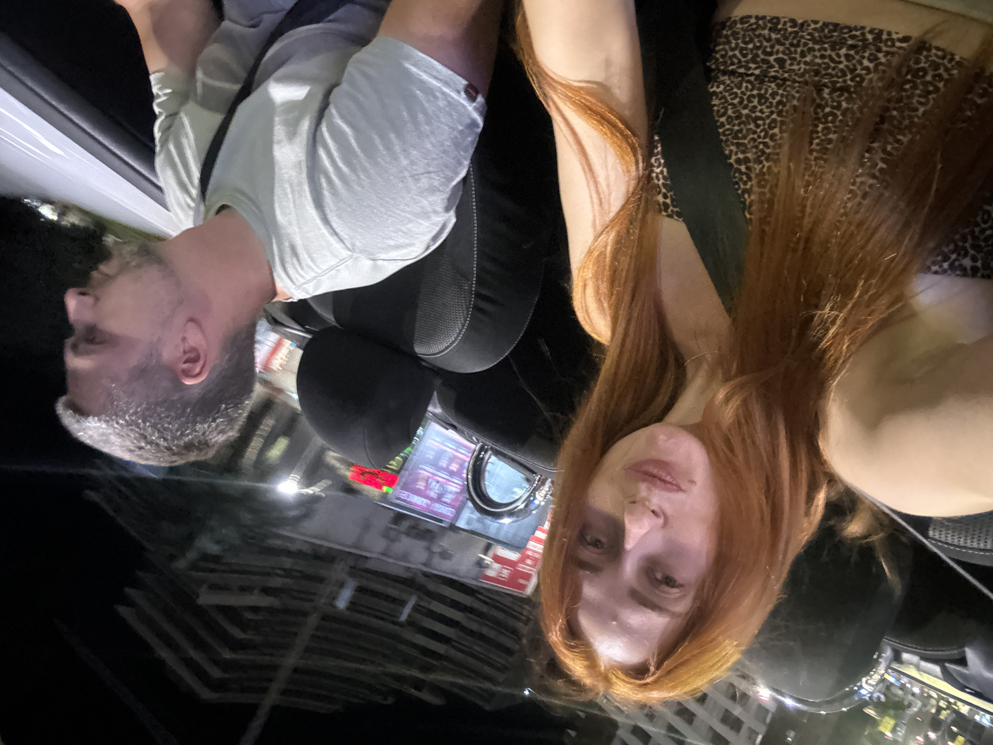 In Budva, Montenegro, two individuals are seated in what appears to be a convertible automobile at night. A woman with long, auburn hair is in the foreground, positioned on the left side of the frame, taking a selfie. She is wearing a leopard-print halter top. Beside her, on the right, is a man with short, graying hair, wearing a light gray t-shirt. He is looking out of the vehicle to his right. The background reveals the exterior of buildings with visible architectural details and lighting, suggesting an urban environment. The interior of the car is dark, with black seats and a seatbelt visible across the woman. The overall lighting conditions indicate it is nighttime, with artificial lights from the surroundings illuminating parts of the scene.
