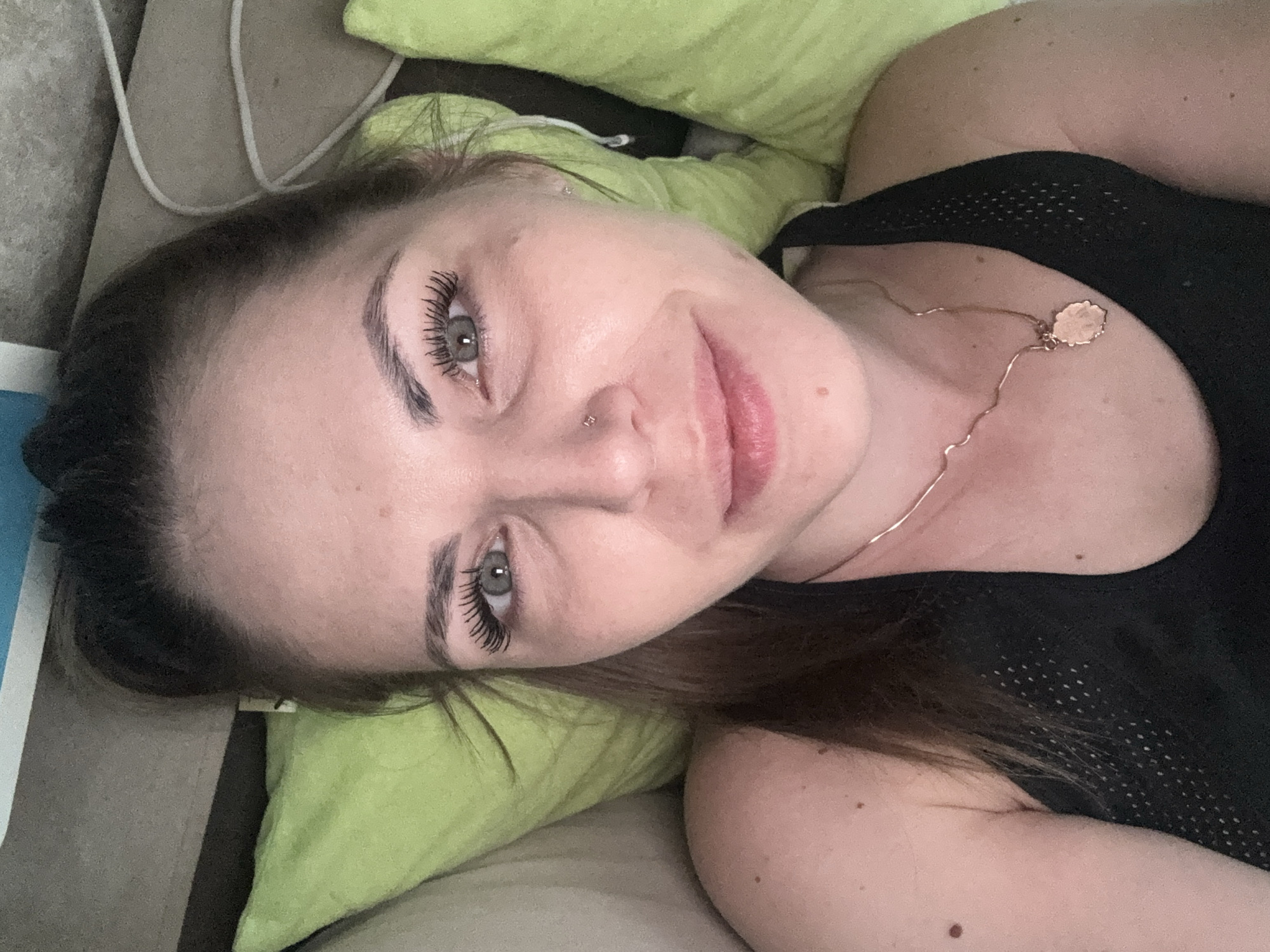 A woman with fair skin, light gray eyes, and brown hair is lying on a light-colored sofa with lime green pillows. She is wearing a black mesh tank top and a thin gold necklace with a round pendant. Her face is turned toward the camera, and she has a slight smile. She has a nose piercing. There is a white cable visible in the upper left corner of the image. The location of the photo is not identifiable as Раздолье, Russia.