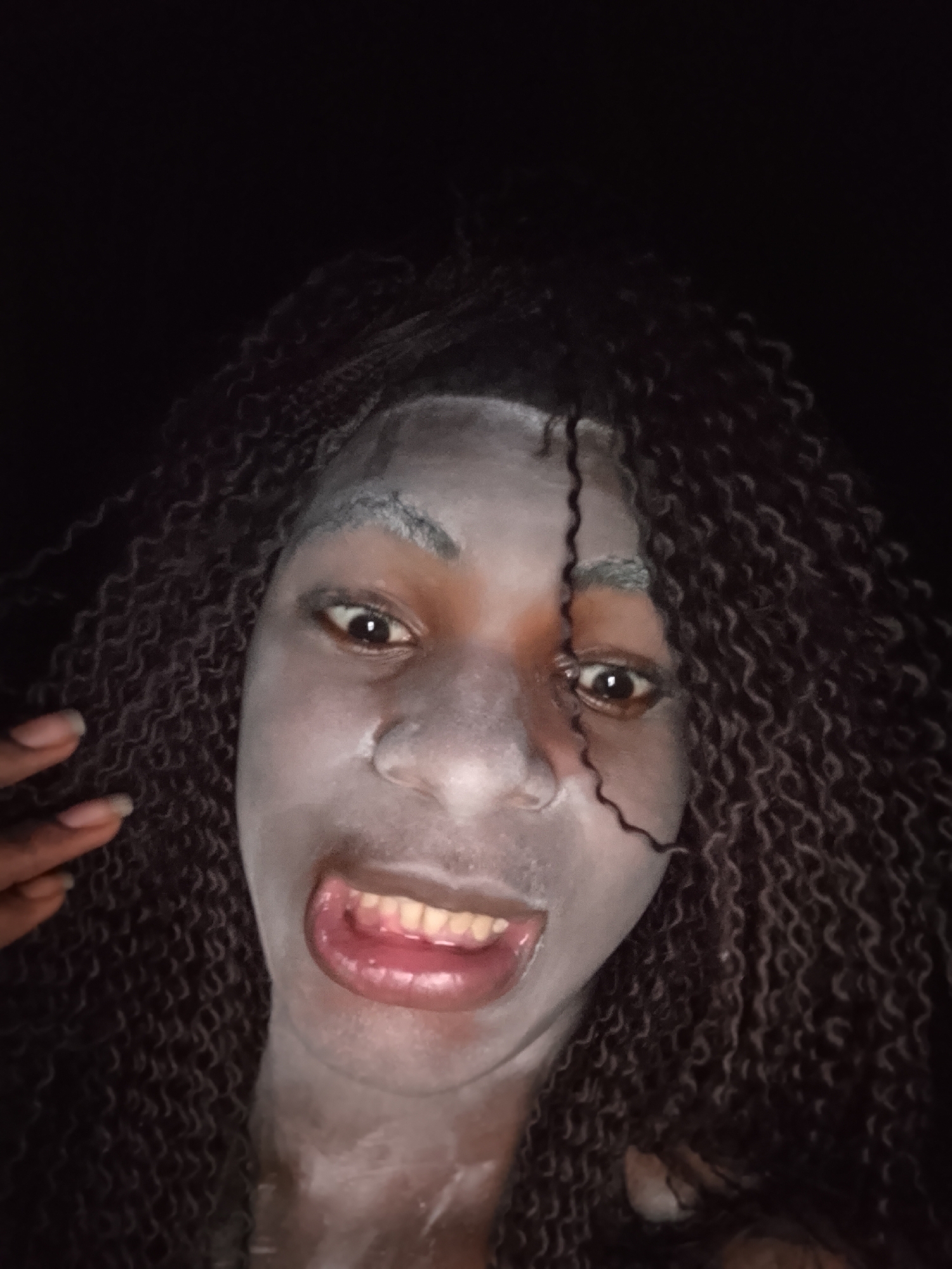 A close-up portrait features a person, appearing to be a woman with dark skin, against a uniformly black background, suggesting a dark environment or night-time setting. Her face and visible parts of her neck and chest are covered in a white substance, resembling powder or paint. She has long, dark, tightly curled hair that frames her face, with some strands falling across her forehead. Her eyes are wide open and dark, staring directly into the camera with an intense or surprised expression. Her mouth is open wide, revealing both upper and lower teeth, as well as her tongue, indicating a loud expression like a yell, gasp, or a theatrical grimace. On the left side of the frame, near her cheek, a hand with manicured fingers painted with dark nail polish is visible. The lighting is bright and direct, possibly from a flash, highlighting the details of her face, hair, and the white substance. There are no other objects, identifiable locations, or visible text in the image. The overall mood is expressive and theatrical.
