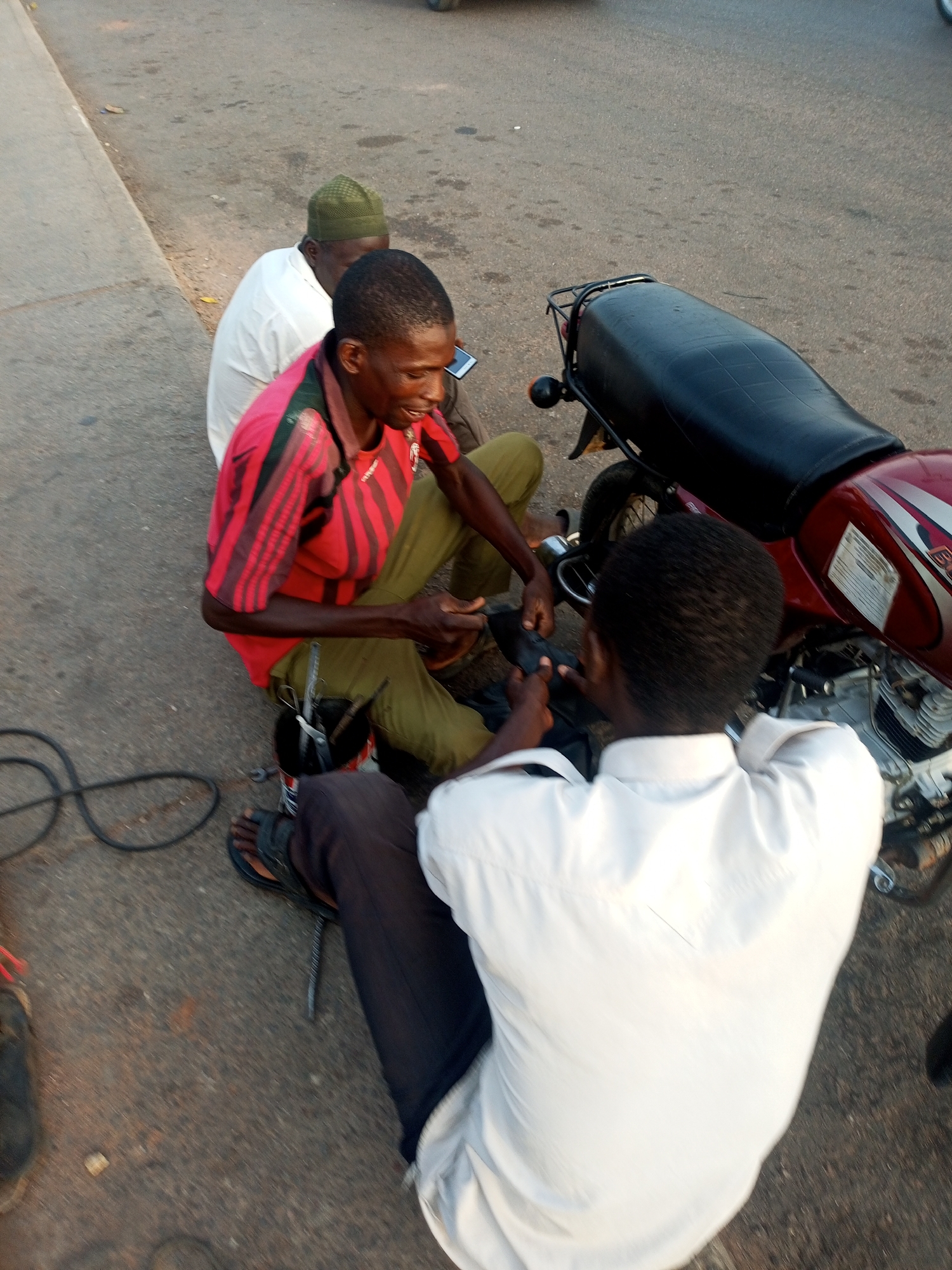 Three men are gathered around a maroon motorcycle on a paved road or street in Unknown, Unknown. One man, wearing a red and black striped shirt and green pants, is crouched and appears to be working on the motorcycle or discussing it with another man in a white shirt who is seated facing the motorcycle. A third man, wearing a white shirt and a green knit cap, sits to the side. Tools and a black cable are visible on the pavement.
