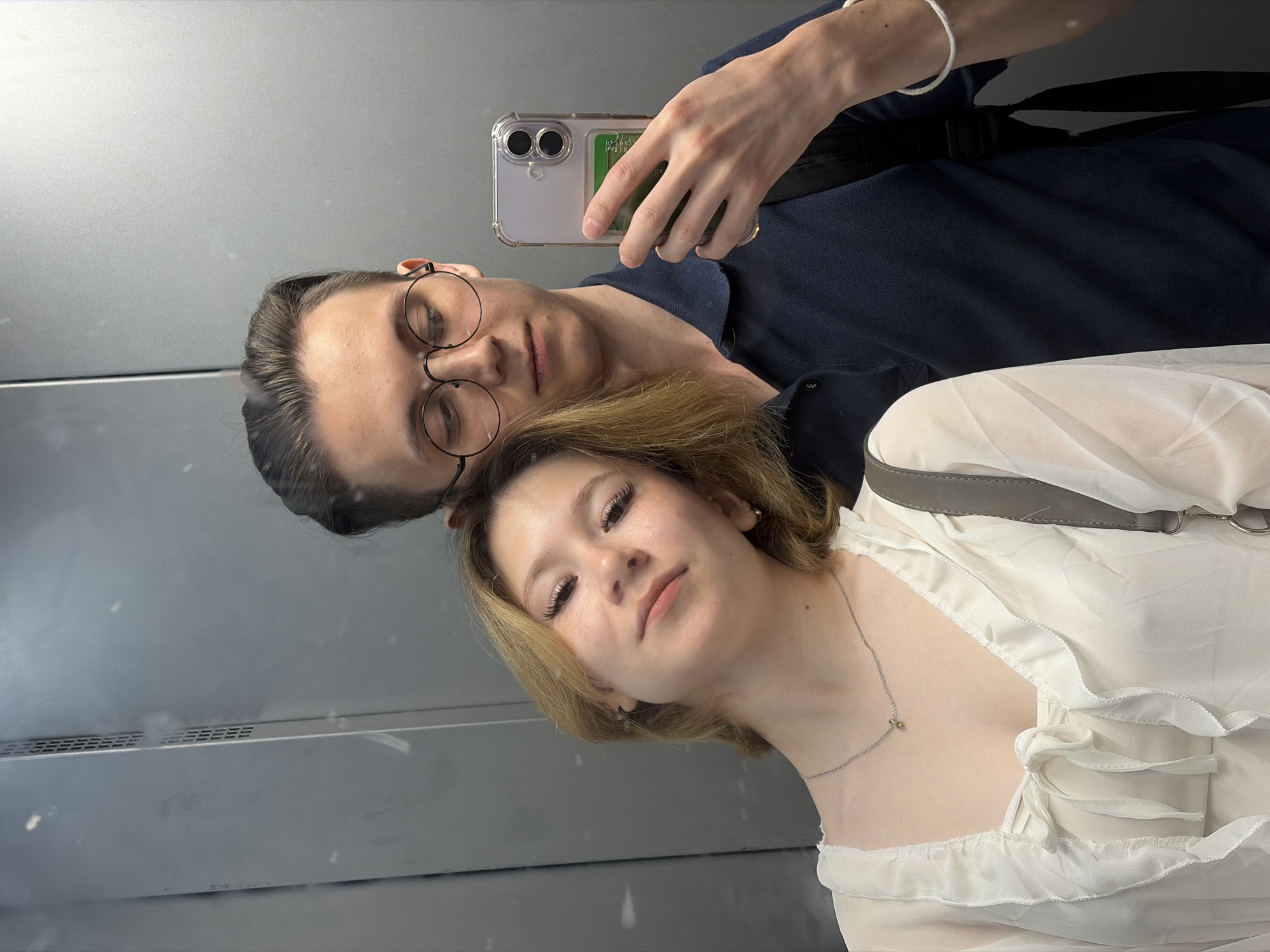 This is a mirrored selfie taken in an elevator. Two young adults are visible, reflected in the elevator doors. On the left of the frame, a person wearing round glasses and a dark blue collared shirt is looking down. On the right, a person with blonde hair and a white ruffled top is looking towards the camera. The person on the right is wearing a delicate silver necklace. The elevator interior appears to be brushed metal, and a ventilation grate is visible in the background. The lighting suggests it is daytime. The overall mood is casual and intimate, as if capturing a shared moment. No visible text is present in the image.