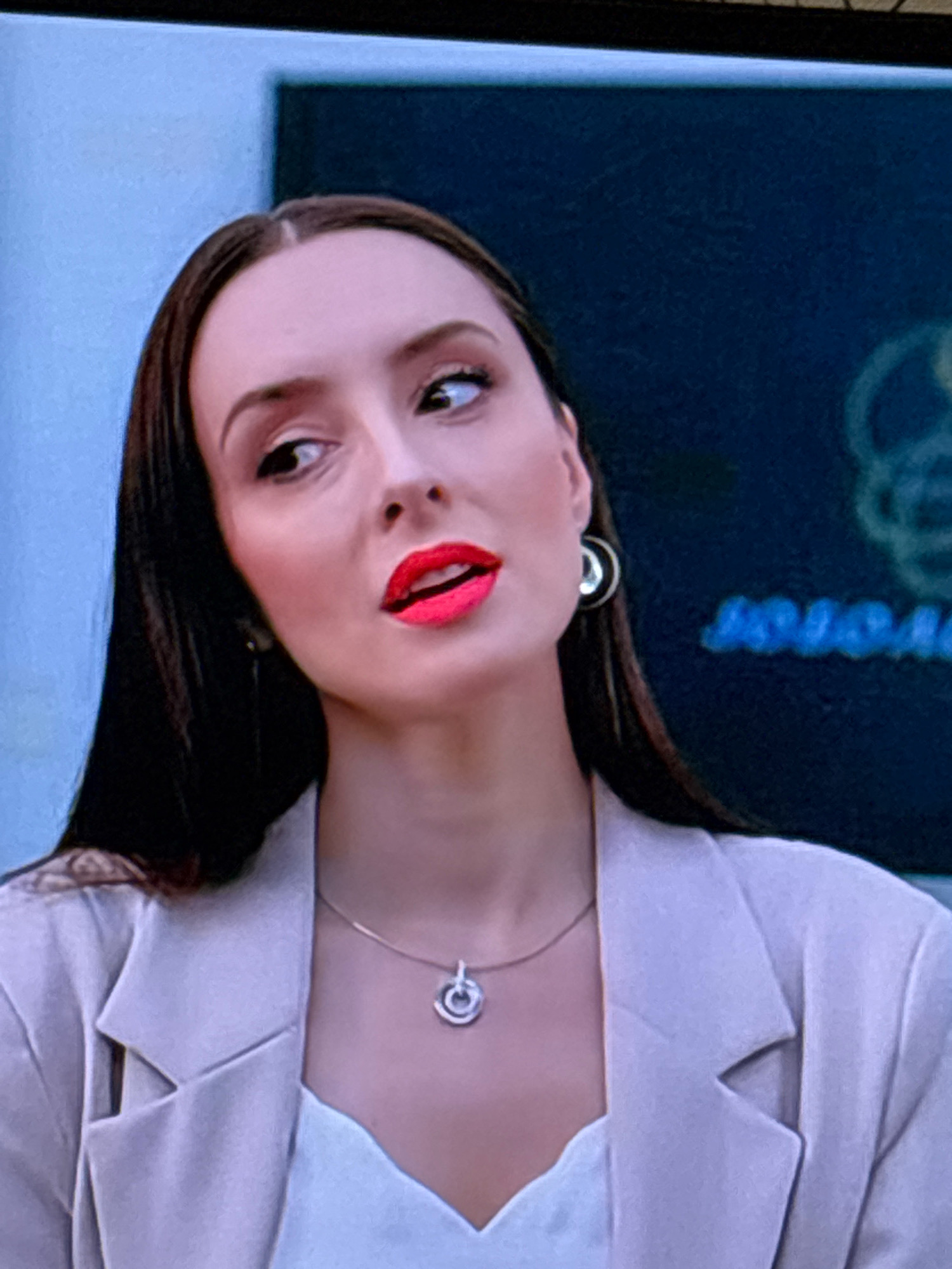 The image is a close-up, slightly blurry, headshot of a young Caucasian woman with dark hair, wearing a light-colored blazer and a white top. She has bright red lipstick on and is wearing a silver necklace with a circular pendant and hoop earrings. She is looking off to her right with her head tilted. The background is a dark blue surface with some lighter blue text or graphics visible, possibly a logo or sign. There is no discernible information in the image that would indicate a location in Sinegorskiy, Russia.