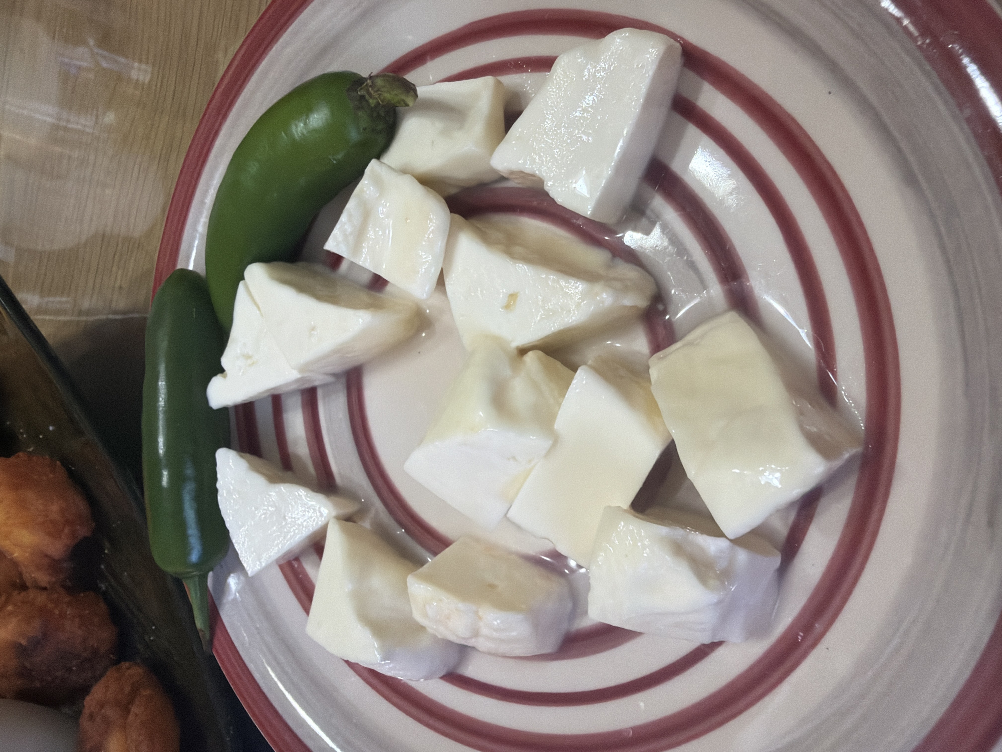 A high-angle, medium-close-up photograph displays a culinary arrangement on a white ceramic plate featuring concentric red spiral stripes. Positioned on the plate are approximately twelve irregularly cut, roughly cubic pieces of white, semi-soft solid food, consistent with fresh cheese or a similar dairy product. Many of these pieces exhibit a glistening, moist surface, suggesting a liquid coating such as brine or oil. Accompanying the cheese are two intact, dark green chili peppers, consistent with the morphology of jalapeños, one positioned near the upper left rim and the other near the upper right rim of the plate. The background, heavily blurred, reveals several golden-brown, irregularly shaped, fried food items resting on a dark surface in the upper left quadrant. The upper right background shows a lighter, vertically grained surface, possibly wood or laminate, also significantly out of focus. The scene, devoid of human subjects or explicit action, captures a static food presentation within an indoor environment in Shumen, Bulgaria.
