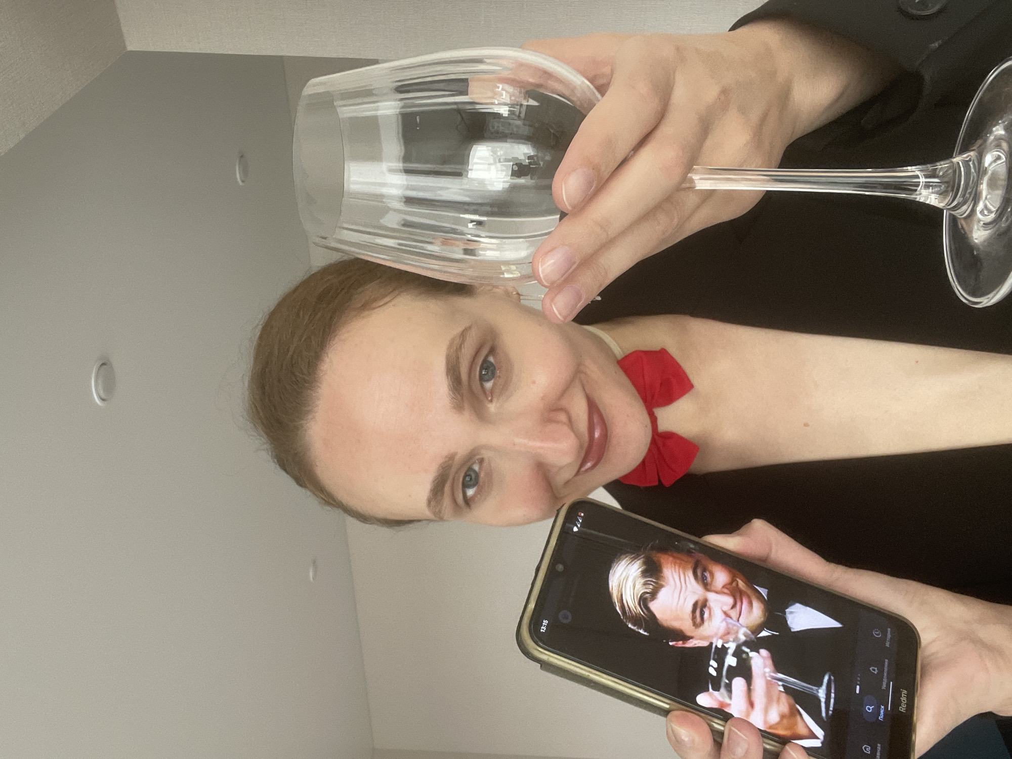 A woman indoors holds a wine glass and a phone displaying an image of Leonardo DiCaprio holding a similar glass. She is wearing a black blazer with a red bow. The setting appears to be inside a building with white walls. There's no visual information to confirm or deny the image being taken in Подшивалово, Russia.