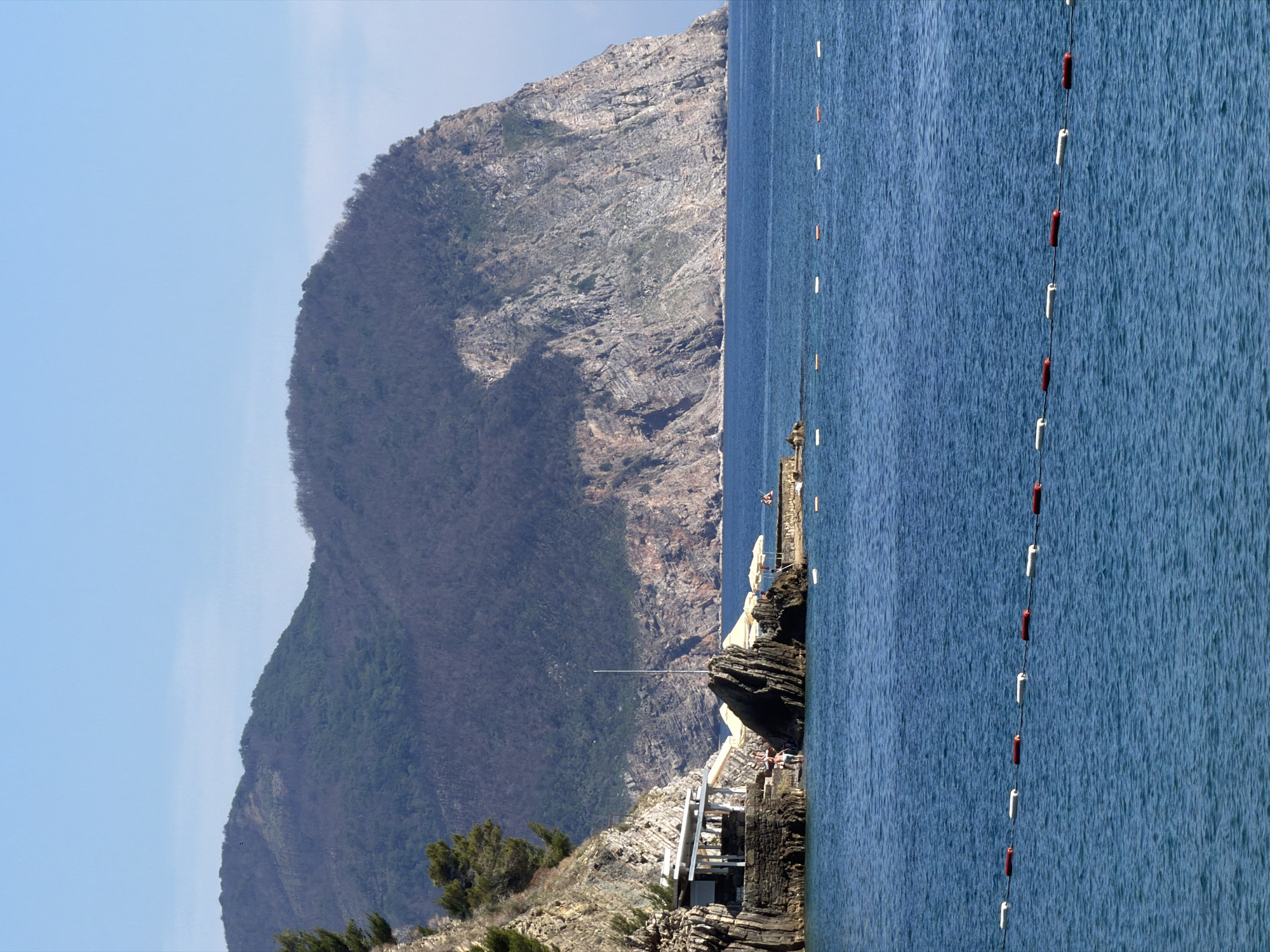 The image depicts a coastal scene in Petrovac centar, Montenegro. In the foreground, the deep blue sea is dotted with a line of red and white buoys. To the left, a rocky shore features a white-roofed structure, possibly a cafe or restaurant, built into the cliff face. Several people are visible near the structure, some sitting under umbrellas. Jagged rock formations rise from the water. In the background, a large, tree-covered mountain slopes down to the sea. The sky is clear and blue.