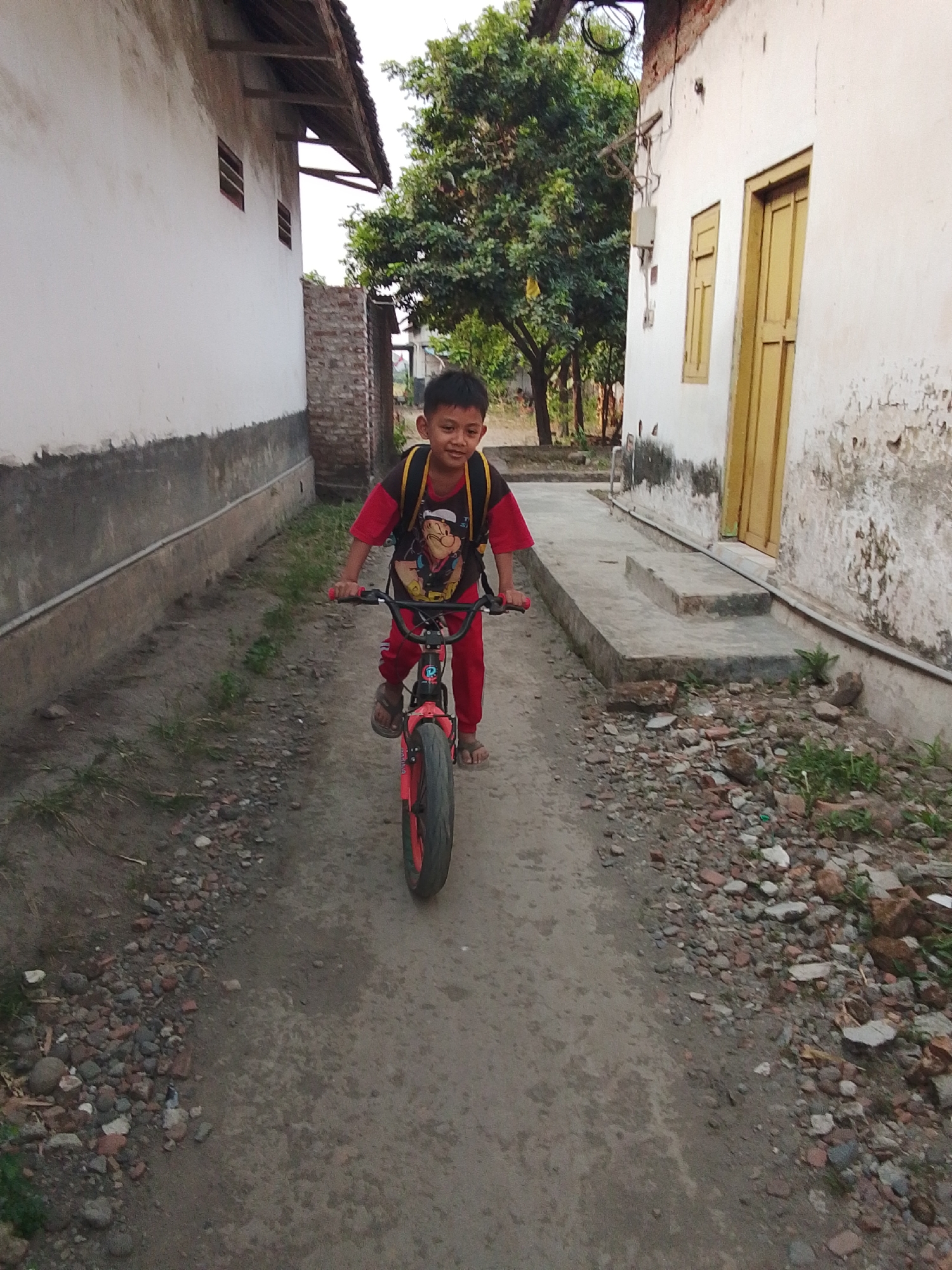A young boy, wearing a red shirt with a graphic print, red pants, and a yellow backpack, rides a red bicycle down a dirt path. He is smiling and looking forward. The path is flanked by white buildings with weathered walls and wooden roofs. Green trees and some debris, including rocks and broken bricks, are visible on either side of the path. The scene appears to be in an outdoor, residential area.