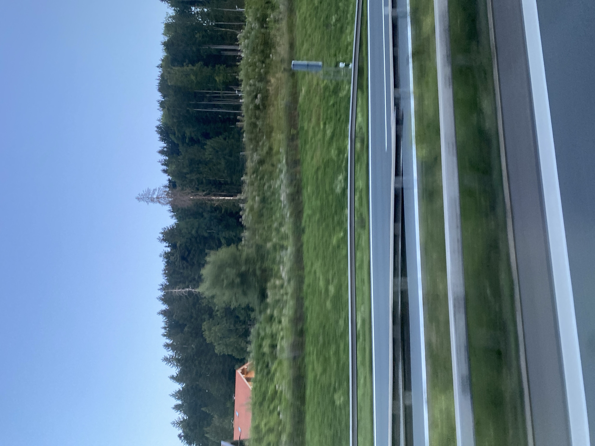 The image depicts a highway scene with a dense forest in the background. A small house with a red roof is visible nestled among the trees on the left side. The sky is clear and blue, suggesting it is either early morning or late evening. The highway itself is bordered by a grassy embankment and metal guardrails. The image is captured from a moving vehicle, indicated by the motion blur. The location is in the municipality of Siegsdorf, Germany, a region known for its scenic beauty and proximity to the Bavarian Alps. The scene is peaceful and serene, devoid of any human presence.