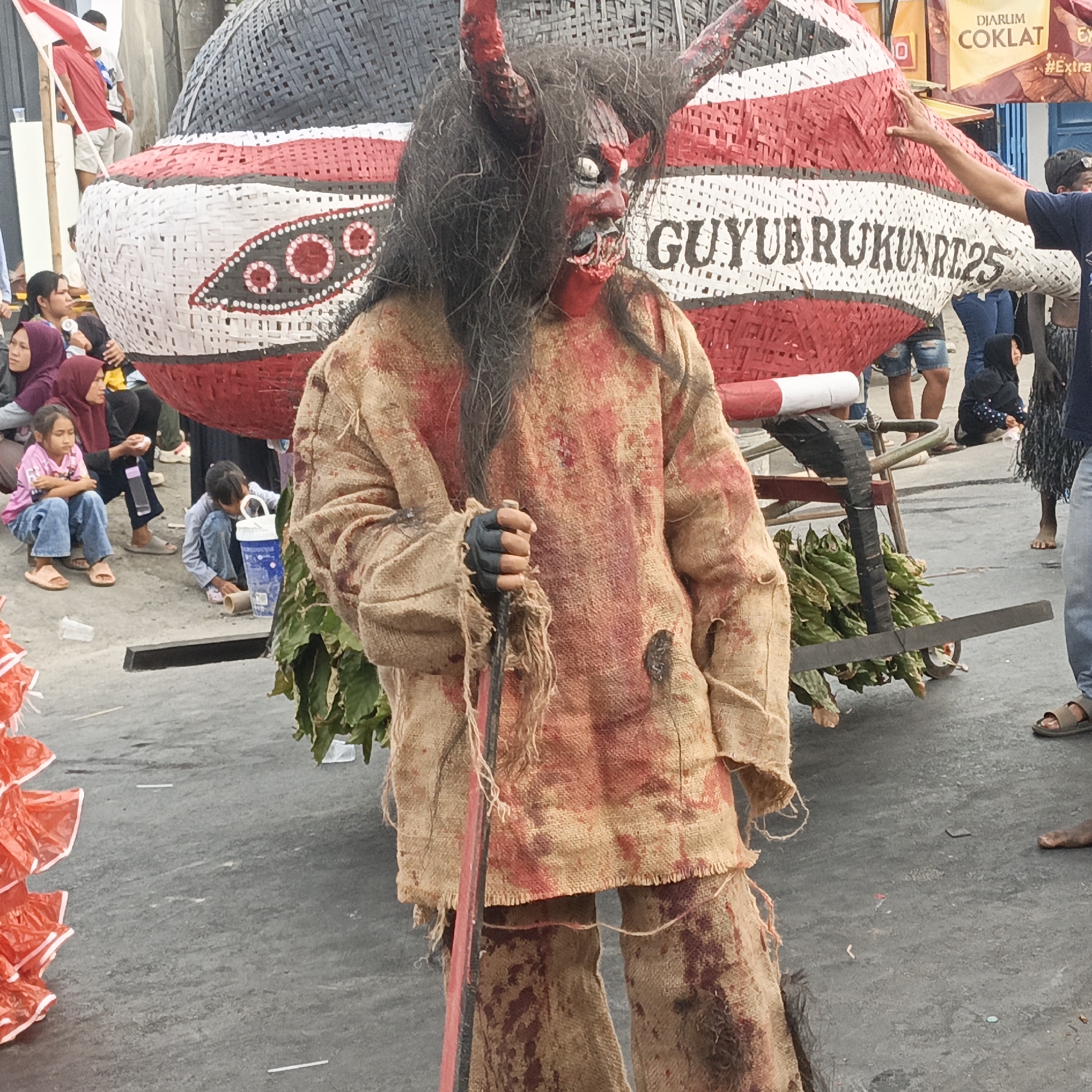 The image depicts a street scene in Randudongkal, Indonesia, likely during a festive or cultural event. In the foreground, a person in a gruesome costume resembling a demon or mythological creature stands prominently. The costume is made of burlap, stained with red to simulate blood, and features dark, messy hair and red horns. The figure holds a dark-colored stick or staff.
Behind the costumed individual, a large, intricately woven structure, possibly a float or part of a parade, is visible. It is decorated with red, white, and black patterns, including circular motifs. Text, "GUYUBRUKUNRT25", is visible on this structure.
Several people are present in the background, including children and adults, some of whom are observing the scene. Some individuals are seated on the ground, while others are standing. A cart laden with green foliage is positioned behind the costumed figure. The ground appears to be paved with asphalt. The overall atmosphere suggests a public gathering or celebration in an urban or semi-urban setting.