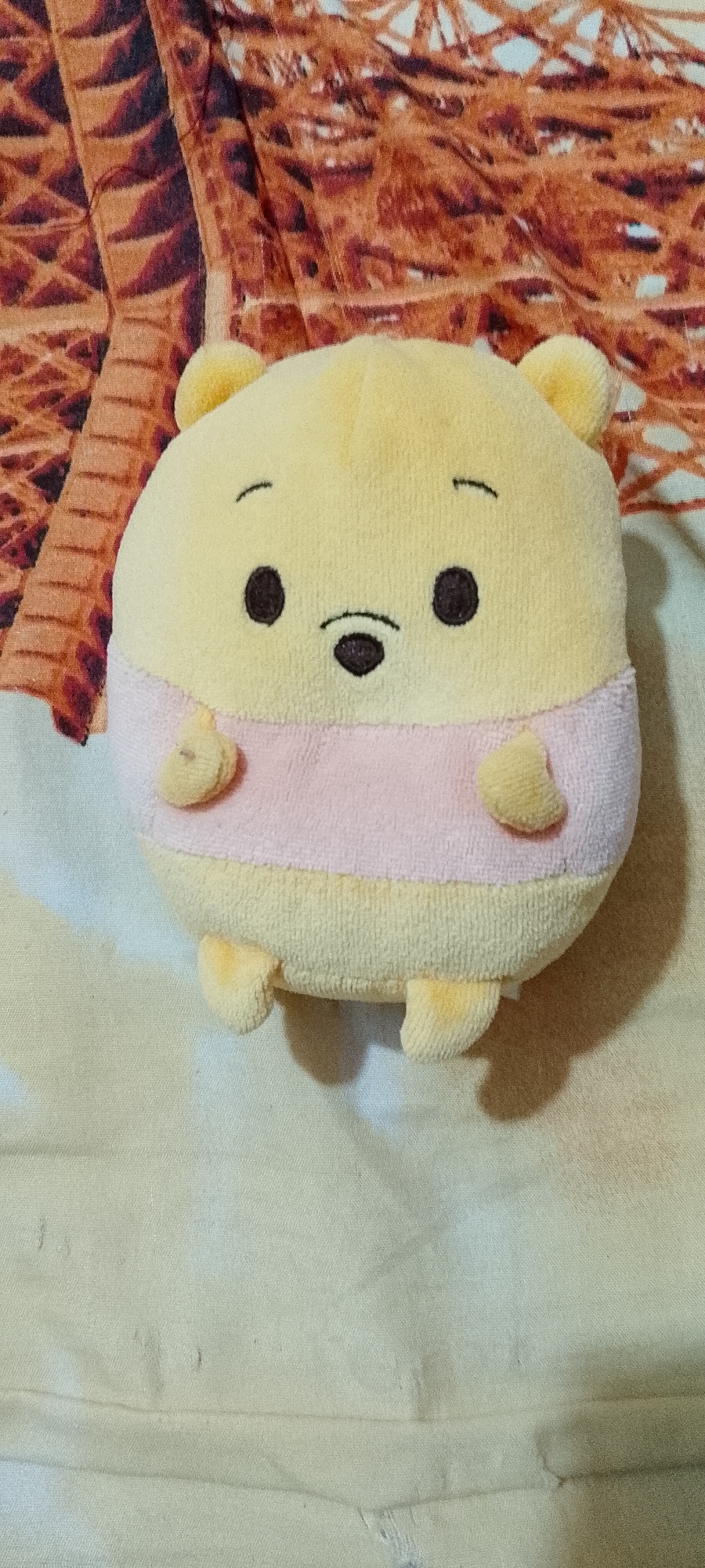 A plush toy, resembling Winnie the Pooh, is positioned on a light-colored surface. The toy is yellow with a pink stripe around its midsection. It has small black eyes, a black nose, and small ears. There are small plush hands and feet visible. The background behind the toy is a patterned fabric with shades of orange and brown. The location is not specified to be in Singkawang, Indonesia.