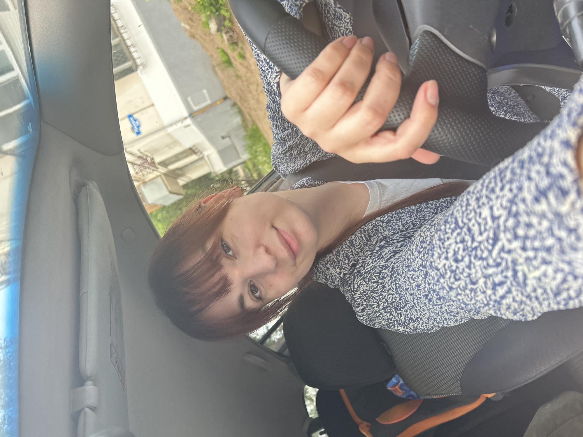 A woman with reddish-brown hair and bangs is seated in the driver's seat of a car, holding the steering wheel. She is wearing a white t-shirt and a blue and white patterned sweater. A sun visor is visible on the left side of the car's interior. Outside the car window, a building and some greenery are partially visible, but the exact location in Chaikovsky, Russia, is not discernible from the image. The interior of the car includes a black seat and what appears to be a child's seat or backpack behind the driver's seat.