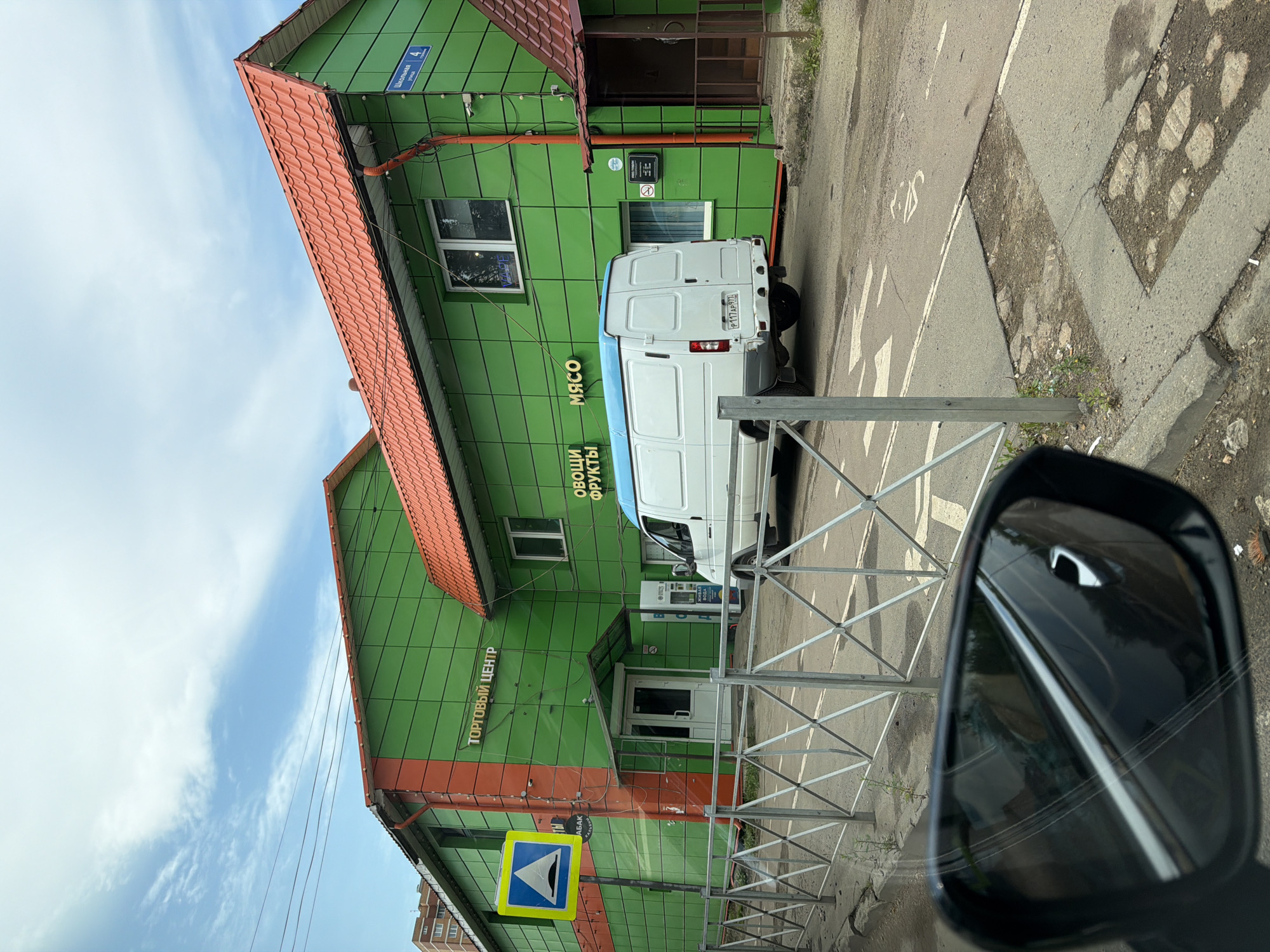 The image shows a commercial building with a green facade and a red roof, likely a "Торговый Центр" (Trading Center). Signs on the building indicate "Овощи" (Vegetables) and "Мясо" (Meat). A white delivery van is parked in front of the building. A yellow and blue traffic sign indicating a speed bump is visible to the left. A metal fence runs along the front of the property. The scene appears to be in a city, with a cloudy sky and some vegetation. The reflection in the car's side mirror shows more of the street and surroundings, including a street sign that reads "Школьная 4" (Shkolnaya 4), indicating the location is likely on Shkolnaya Street, building number 4, in Povarovo, Russia. The ground is asphalt with some cracks and patches, and there is evidence of litter. There are no people visible in the image.