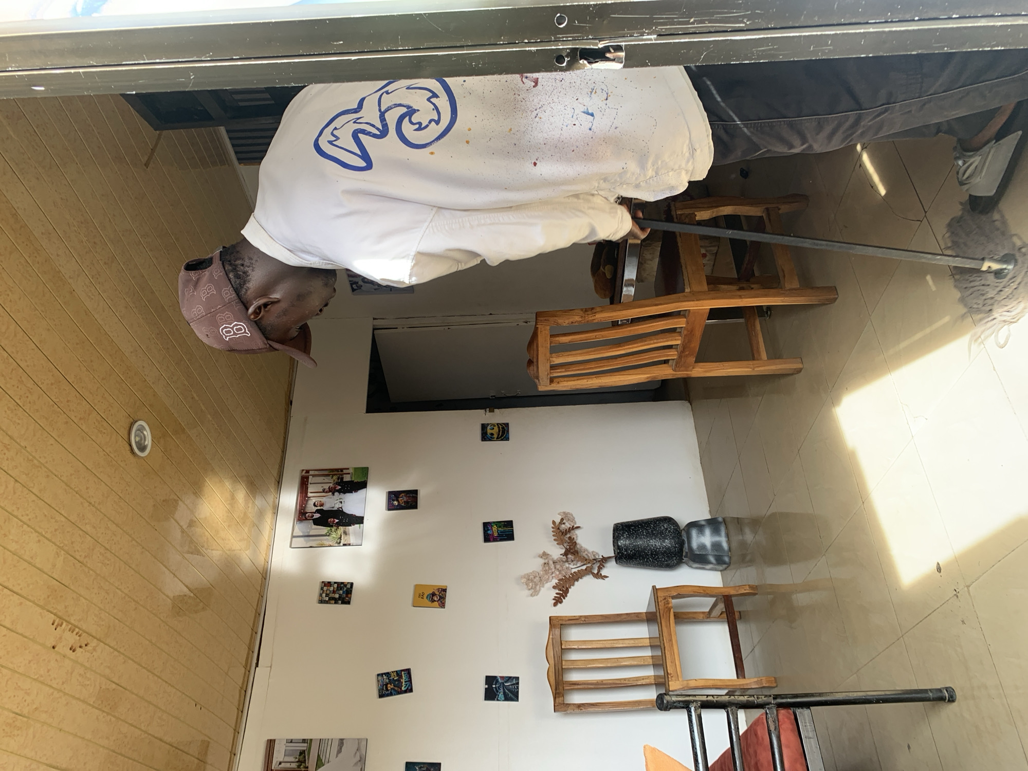 The image, taken in Jos, Nigeria, depicts an indoor scene where a dark-skinned man is mopping a light-colored tiled floor. He wears a brown baseball cap with a white "B" logo (similar to the Boston Red Sox emblem), a white long-sleeved shirt with a large blue swirled graphic (resembling a stylized '3' or 'E' or a sports logo) and scattered paint splatters, and dark trousers with white sneakers. He is actively cleaning with a black-handled mop with a grey string head.
The room features a light brown wooden paneled wall on the left side, with a circular ceiling light fixture embedded in it. The right wall is white and decorated with numerous small framed pictures or postcards, and one larger framed photograph showing several people. Text visible on some of the small pictures includes "BATMAN" and "FUNK." A black vase holding dried decorative plants and several light wooden stools or low chairs are arranged against the white wall. Another metal-framed piece of furniture is partially visible in the foreground. Bright sunlight streams into the room, creating distinct shadows on the tiled floor, indicating it is daytime.