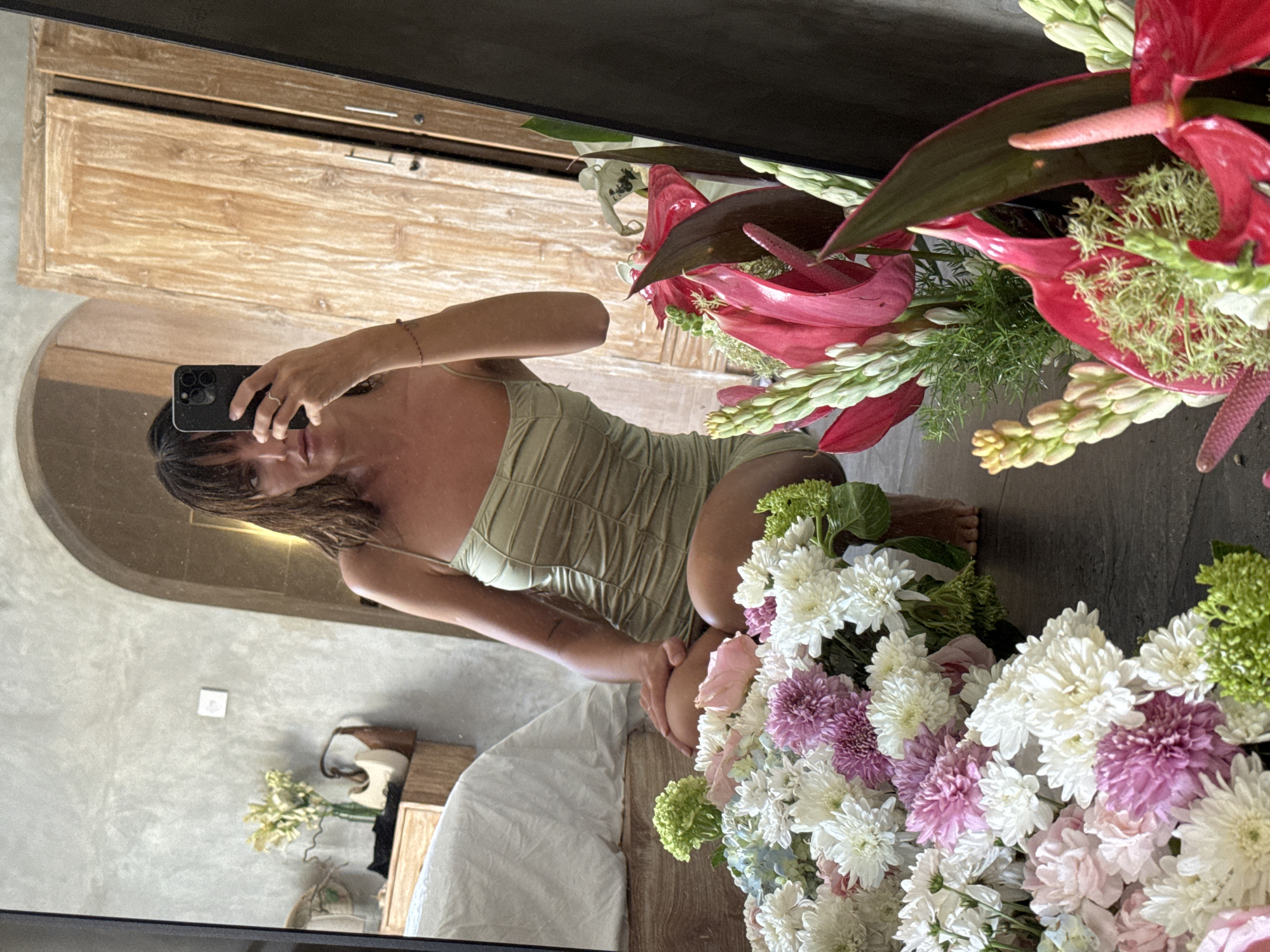 A mirror selfie captures a woman in a green dress kneeling on a bed or floor next to a large bouquet of flowers. The mirror reflects a bedroom with a bed and a wooden cabinet. The bouquet consists of pink, white, and green flowers. The woman is holding her phone to take the picture. The location is identified as North Kuta, Indonesia, based on architectural and decorative elements consistent with the region.