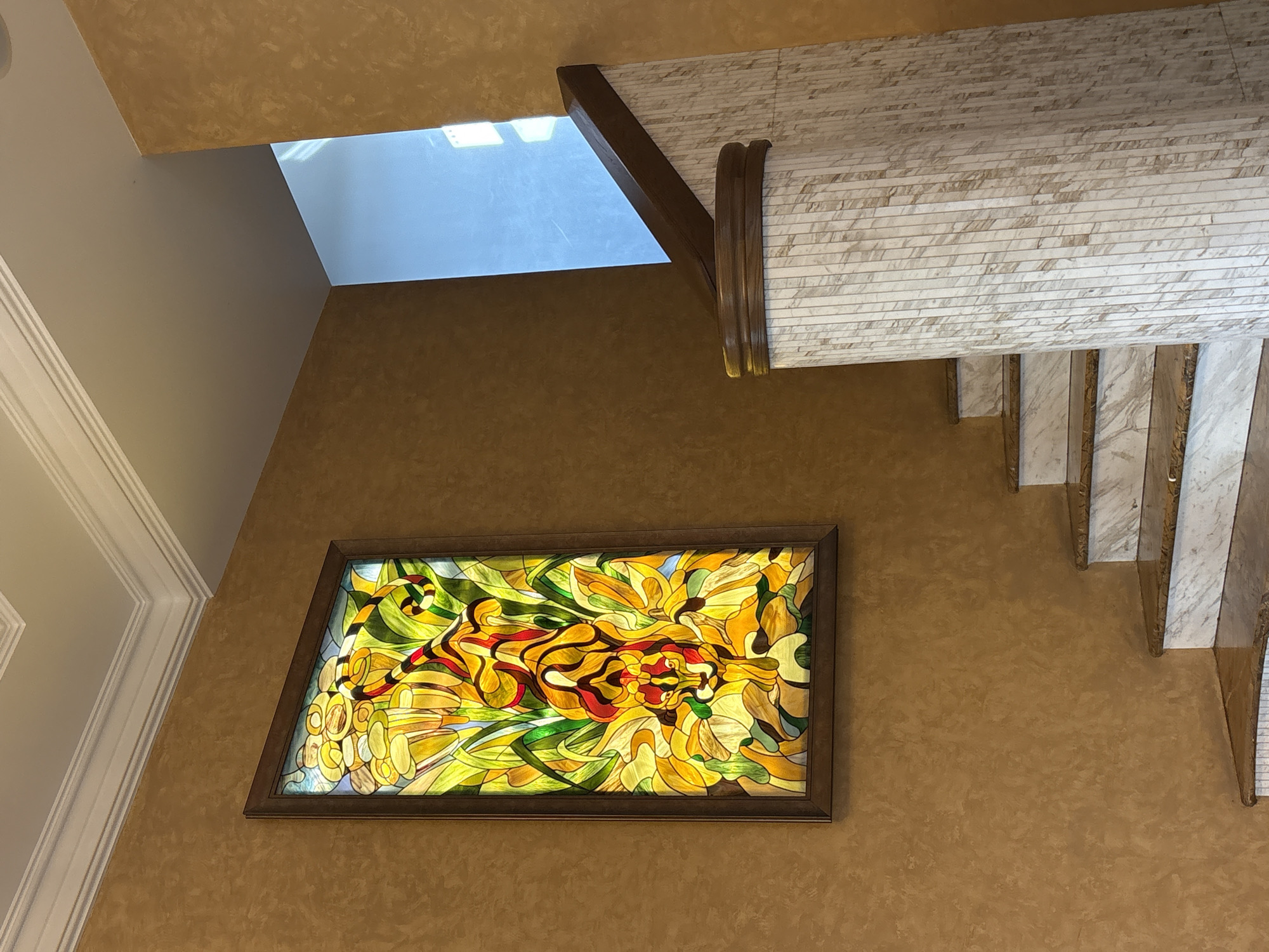 The image shows an interior view of a residential hallway, possibly in Khabarovsk, Russia. The focal point is a stained-glass window with a horizontal orientation, framed in dark wood, depicting an abstract or stylized design in vibrant yellows, reds, and greens. The window is mounted on a textured brown wall.
Above the stained-glass window, a portion of a skylight is visible, allowing natural light into the space, suggesting it is daytime. To the right of the skylight, a section of a staircase is visible, characterized by marble-like treads and risers, accented with polished brass trim. A wooden beam supports the ceiling structure, which is clad in the same marble-like material.
To the left of the stained-glass window, a section of ornate white crown molding is visible against a lighter-colored wall, indicating a more traditional architectural element in the interior design. The overall impression is one of a well-decorated home, with a mix of modern and classic design elements. No people are visible in the image, and no text is present. The lighting suggests it is daytime, and the weather outside cannot be determined.