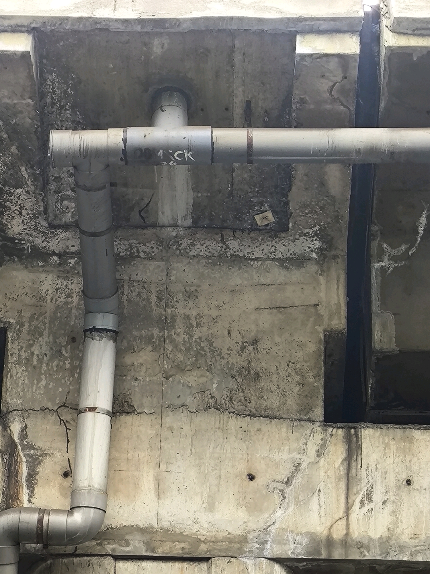 A close-up, eye-level, static medium shot captures a series of grey PVC pipes against a rough, textured concrete background. The pipes are arranged in a T-junction, with one horizontal pipe extending across the frame and a vertical pipe descending from it. The horizontal pipe is marked with black lettering that appears to read "20 C K". The concrete surface is weathered, showing signs of age, discoloration, and some grime, particularly where the pipes are attached. The overall impression is one of an external utility or drainage system, possibly located in an urban setting given the construction materials. The lighting is diffused, suggesting an overcast day or an area with limited direct sunlight. There are no people or animals visible in the frame, and the focus is solely on the infrastructure. The scene has a utilitarian and somewhat aged aesthetic, hinting at the functional purpose of the pipes within a larger building or structure.