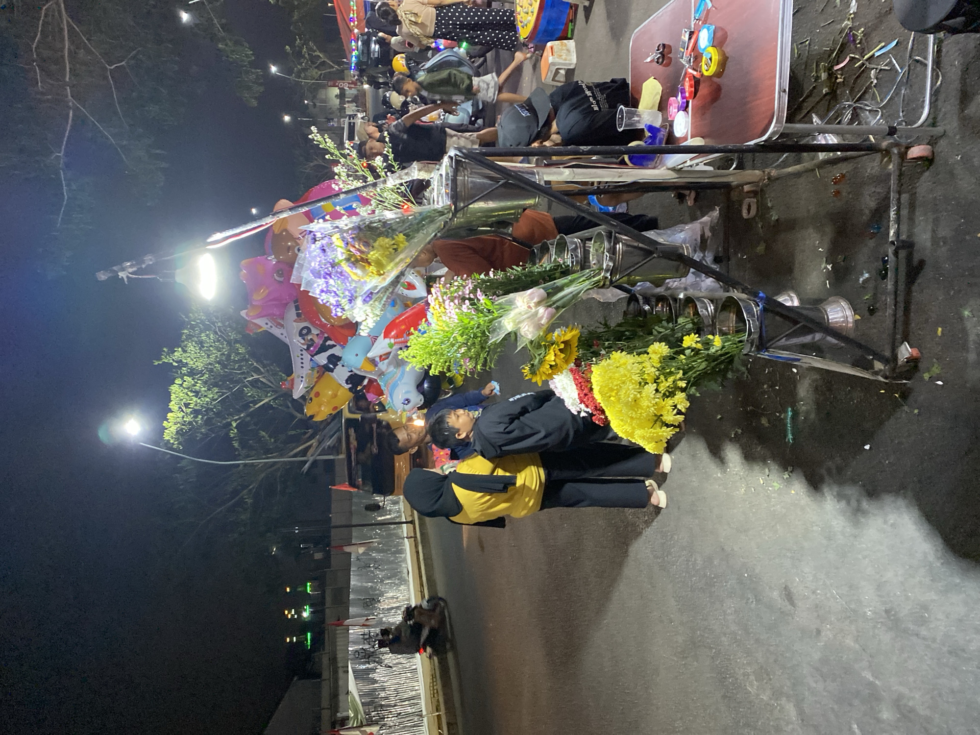 At night in South Tangerang, Indonesia, a street vendor is selling balloons and flowers. The vendor's stall is set up on the side of a road. In the foreground, there are several bouquets of flowers, including yellow chrysanthemums. Behind the flowers, a rack of metallic vases holds more floral arrangements. A group of people, including a woman in a yellow shirt and headscarf, a young boy in a black hoodie, and a man, are looking at the flowers. In the background, a streetlight illuminates the scene, casting shadows on the asphalt. Further down the road, other vendors are visible, with more people and vehicles, suggesting a lively night market atmosphere. A motorcycle rider is seen in the distance on the left side of the image.