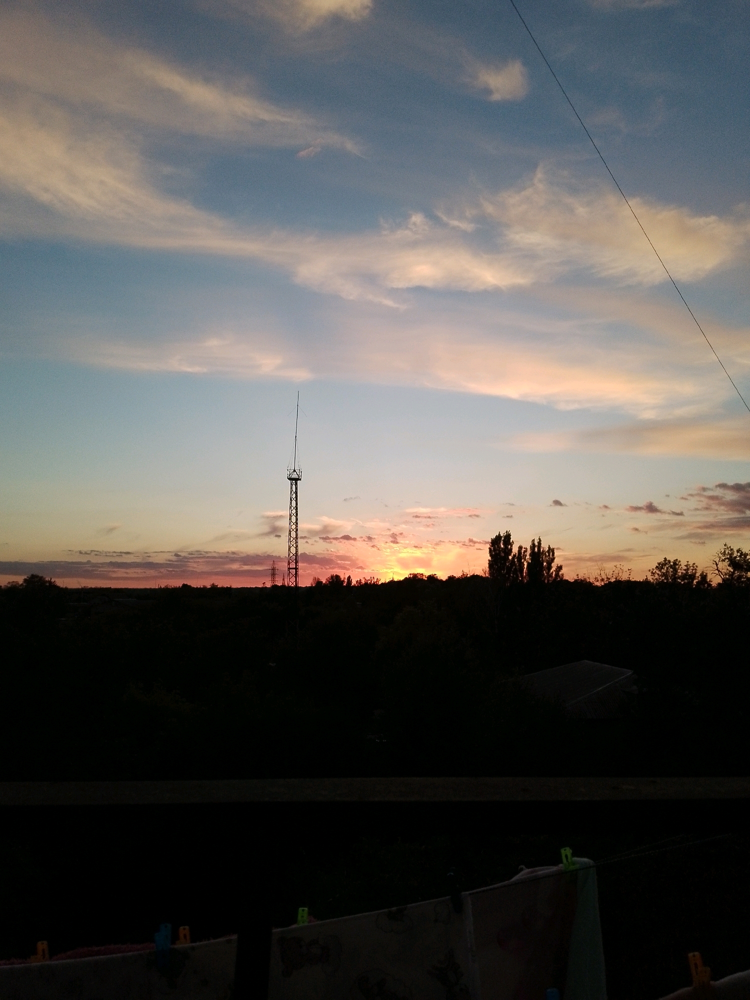 A sunset is visible over a dark, silhouetted landscape. In the center of the image, a tall, lattice radio tower stands against the sky. The sky is a gradient of pale blue near the top, transitioning to soft pinks and oranges near the horizon, with wispy clouds scattered throughout. A thin, dark wire stretches diagonally across the upper right portion of the frame. The foreground is a dark, indistinct area with what appears to be laundry hanging on a line, secured by colored clothespins. The overall scene suggests an outdoor view at dusk.