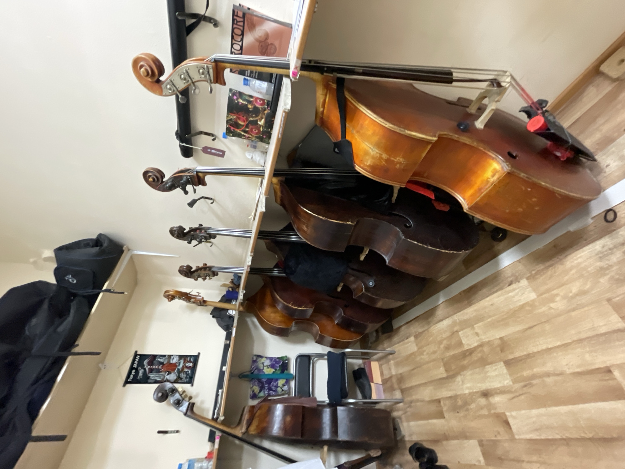 The image shows a storage shelf in a room with wood-patterned flooring. There are five upright basses stored vertically in the shelf. The basses are arranged from top to bottom, with only the top two basses fully visible. A black bag and a poster are visible to the left of the basses on shelves. Other items such as pillows, bottles, and musical instrument accessories are also visible. The room's walls are light in color. It is not possible to determine if the location is in the city of Yekaterinburg, Russia.