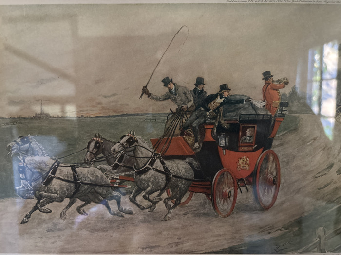The image is a photograph of a framed historical print, depicting a four-horse mail coach in rapid motion across an open landscape. Reflections on the print's surface indicate it is displayed under glass.
The central subject is a vibrant red and black four-wheeled coach, angled from the lower-left foreground, moving towards the right background. It is drawn by four light-colored (grey or white) horses, each equipped with dark harnesses, shown mid-gallop, conveying speed and dynamism.
Five individuals are prominently visible on the coach. The driver, seated on the elevated front bench, wears a dark top hat and light jacket, holding reins and a long whip raised in his right hand. Two additional male passengers in dark top hats and coats are seated next to him. At the rear of the coach's upper deck, a man in a bright orange-red jacket and dark top hat holds a long brass horn to his mouth. Through a small, dark-framed window on the coach's side, a partial view of a passenger is discernible. The side of the coach bears a gold crest and the word "LONDON" in white lettering on a dark panel.
The environment features a dirt or unpaved road, showing tire tracks and implied movement. The background to the left comprises flat green fields extending to a distant horizon, where a cluster of buildings, including a tall smokestack or tower, is visible. The right side of the road is bordered by sparse foliage and a faint wooden fence line. The sky is overcast, displaying a gradient of grey and pale brown tones.
Text in the upper-right of the print identifies the publisher as "Raphael Tuck & Sons Ltd. London, New York. Publishers to Her Majesty the Queen." A partial artist's signature is present in the lower right. The depicted scene, a historical four-horse mail coach, is identifiable as being set in Great Britain due to the inscription "LONDON" on the coach and the British publisher's details. This particular print, as photographed, is located in the city of Monterey, United States of America.