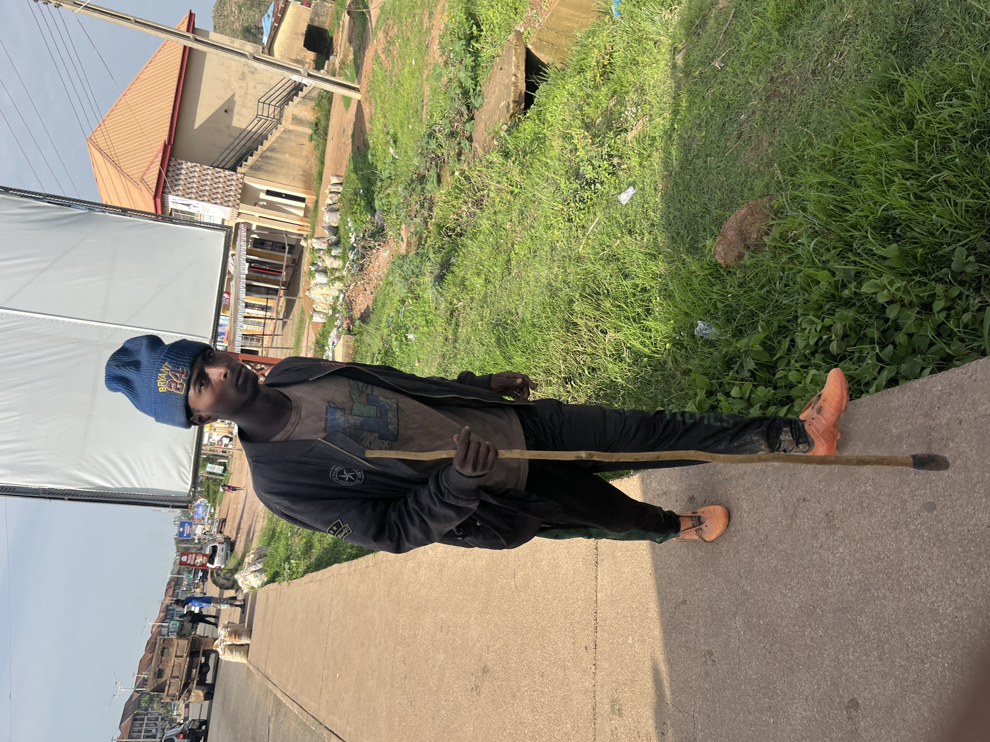 A young African male wearing a blue beanie and a dark jacket over a printed t-shirt is walking on a concrete sidewalk, holding a wooden walking stick. He is also wearing black pants with a green stripe and orange sneakers. The scene appears to be in an urban area, possibly Gura Top, Nigeria, with a road to the left and a grassy area with some debris and a small concrete culvert to the right. Buildings and other vehicles are visible in the background. The sky is clear and bright.