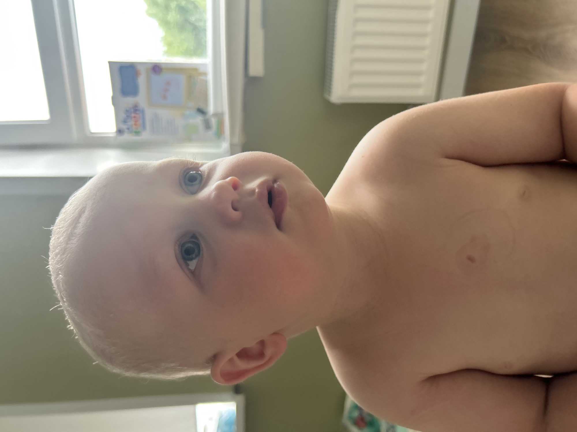 A close-up, eye-level, medium shot captures a toddler, likely male, from the chest up, facing right and looking away from the camera. The child has fair skin, blond hair, and blue eyes. The background features a window with natural light streaming in, a white radiator, and a muted green wall. A colorful children's educational board is visible on the windowsill. The scene appears to be indoors, possibly a child's room or a common area in a home, suggesting a domestic setting in Kaluga, Russia.