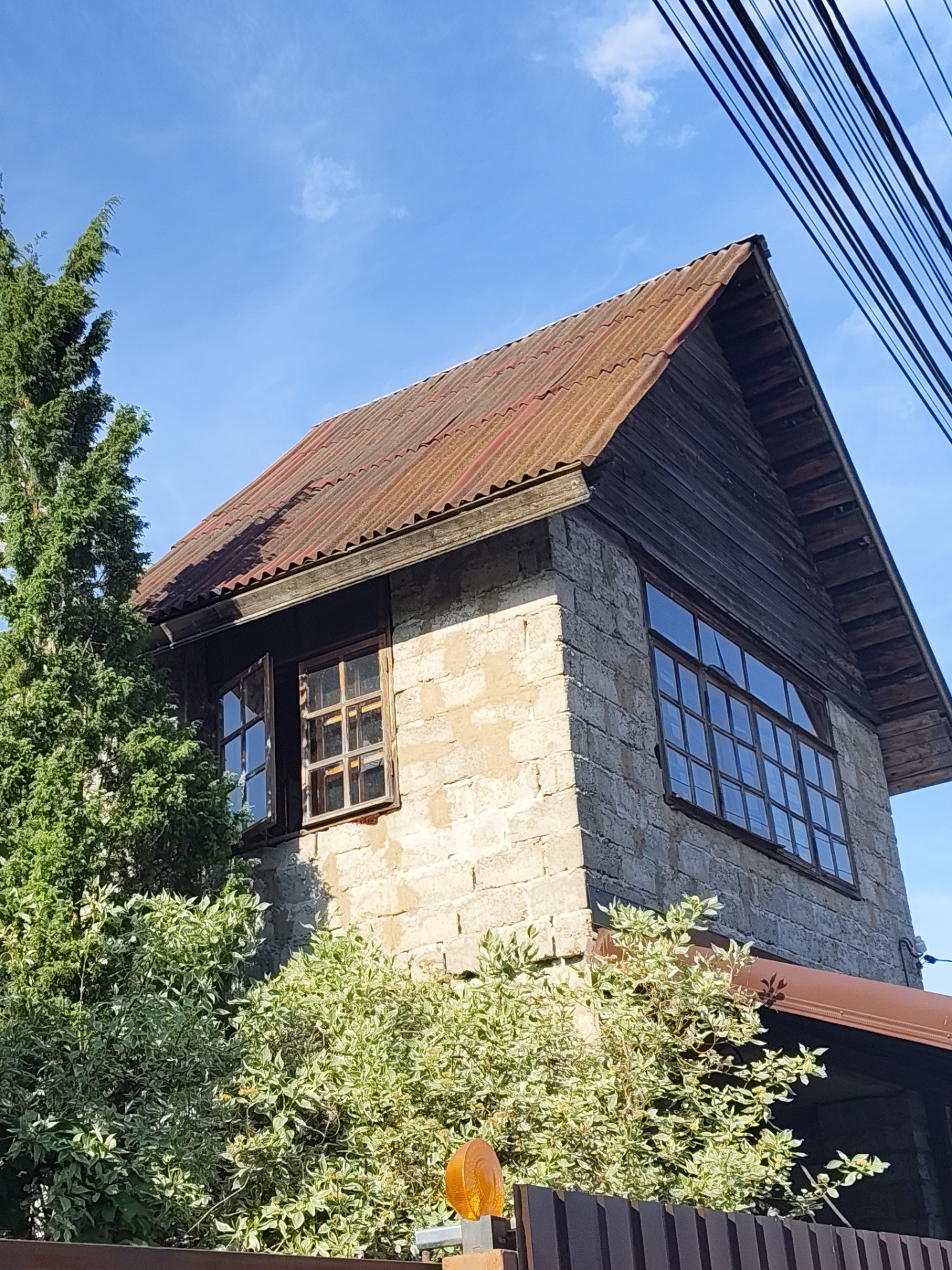 The image depicts a two-story building located in Быково, Russia, viewed from a low angle on a clear day with a bright blue sky and minimal wispy clouds. The building's walls are constructed from light-toned, possibly concrete or cinder blocks with visible mortar lines, showing signs of weathering. A steep gable roof, covered with corrugated reddish-brown metal sheets, exhibits extensive rust and oxidation. The gable ends are clad with dark horizontal wooden planks.
On the building's left side, two windows with dark wooden frames and multiple panes are present; the leftmost window is open outwards. On the right side, a larger, multi-paned window with a dark wooden frame is visible. A dark brown rain gutter or awning extends below the large window.
In the left foreground, a tall evergreen tree with dense green foliage partially obscures the building. In the lower midground and foreground, variegated green and white bushes are prominent, partially concealing a dark brown corrugated metal fence. An orange, dome-shaped light fixture is mounted on a post in the bottom right foreground. Several dark, parallel utility lines or cables diagonally traverse the upper right quadrant of the sky. Bright sunlight illuminates the scene, casting shadows consistent with a clear day.