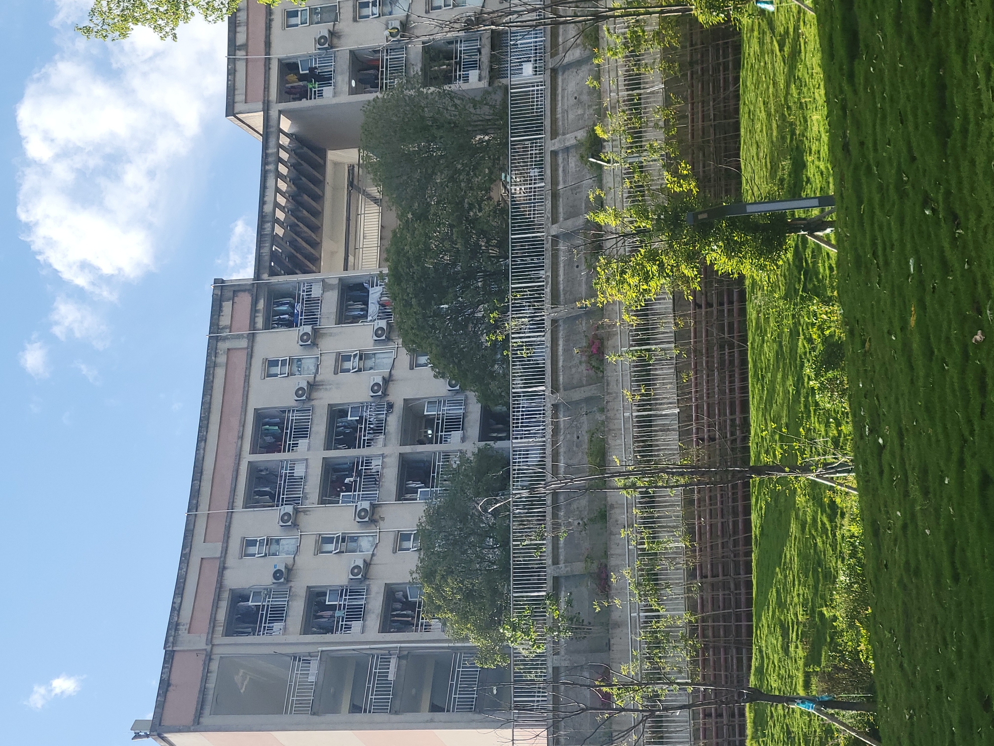 The image shows a multi-story apartment building in Beibei, China. The building is made of concrete and has rows of windows, many of which have air conditioning units installed. Clothes are hanging out to dry on some of the balconies. In front of the building is a strip of green lawn with young trees planted along a metal fence. The sky is visible with scattered clouds. The perspective is from the ground, looking up at the building.