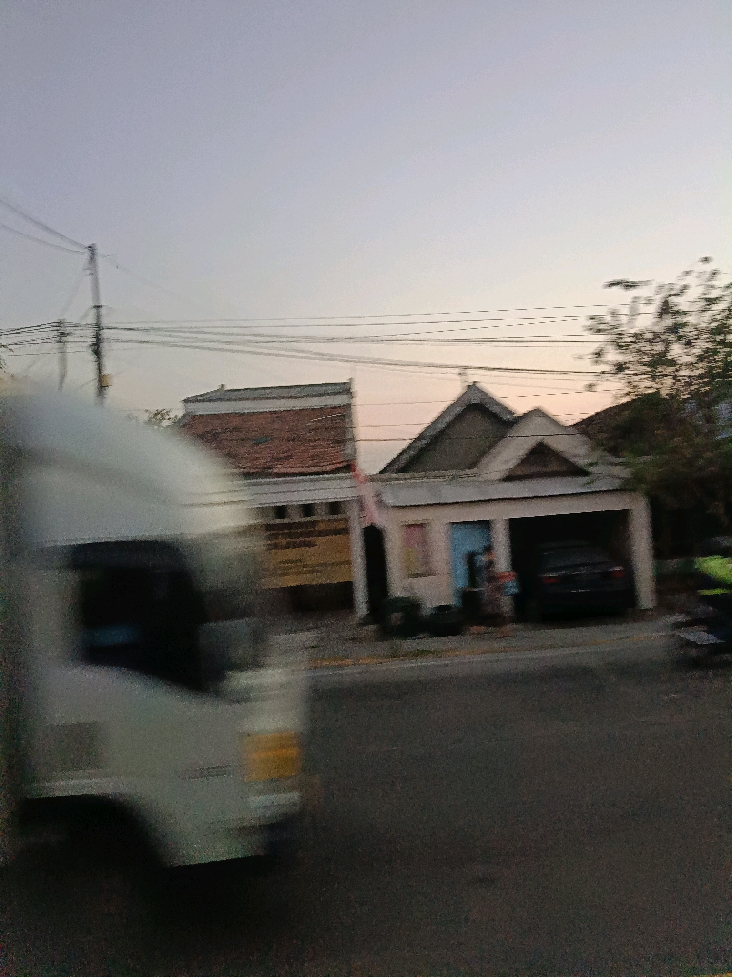 The image captures a street scene in Sumari, Indonesia, during twilight. A white vehicle, blurred due to motion, occupies the left foreground. In the background, two buildings are visible. The building on the left features a red-tiled roof and what appears to be a yellow sign. To its right, a smaller building has a grey, peaked roofline above an open garage area where a dark-colored car is parked. A person is standing near the entrance of this second building. Utility poles and power lines are present in the sky. The sky itself is a pale gradient, indicative of the early evening.