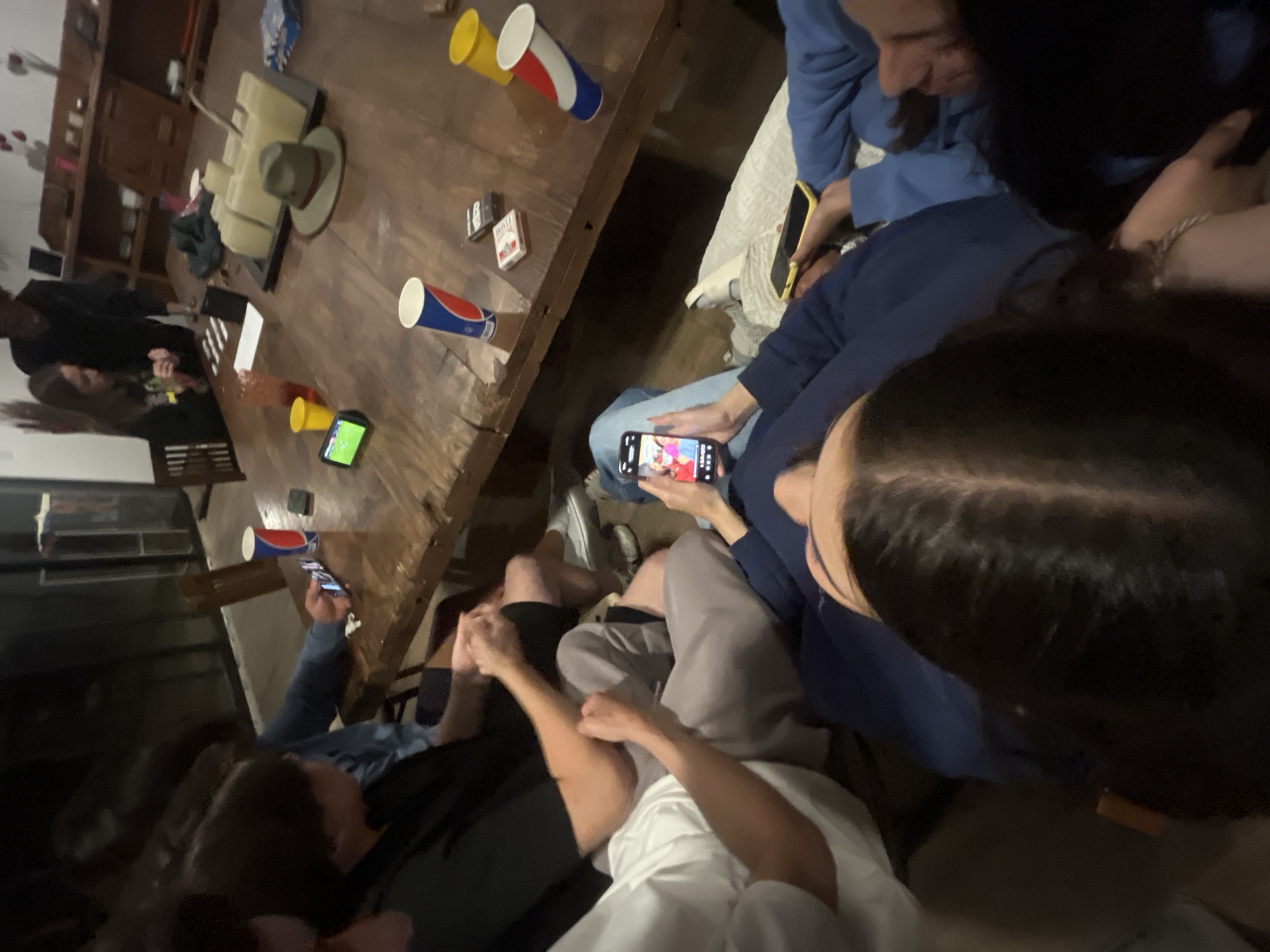 **Scene:** Indoor gathering, possibly a residence, in El Oasis Valsequillo, Mexico.
**People:** Group of individuals (approximately 8-10) seated closely. Predominantly young adults. Some are looking at phones.
**Subjects:**
* Individuals: Wearing casual attire (sweaters, t-shirts).
**Objects:**
* Large, rectangular wooden table: Central focus of the scene. Various objects on the table:
* Disposable cups (red/white/blue, yellow).
* Deck of cards.
* Hat.
* Cell phones (various models).
* Assorted furniture (sofa, chairs).
**Actions/Interactions:**
* Individuals are gathered around the table.
* Some are holding and looking at cell phones.
* Appears to be a social gathering.
* One person is showing their phone screen to others.
**Environment:**
* Dimly lit room.
* Residential setting.
* Informal and relaxed atmosphere.