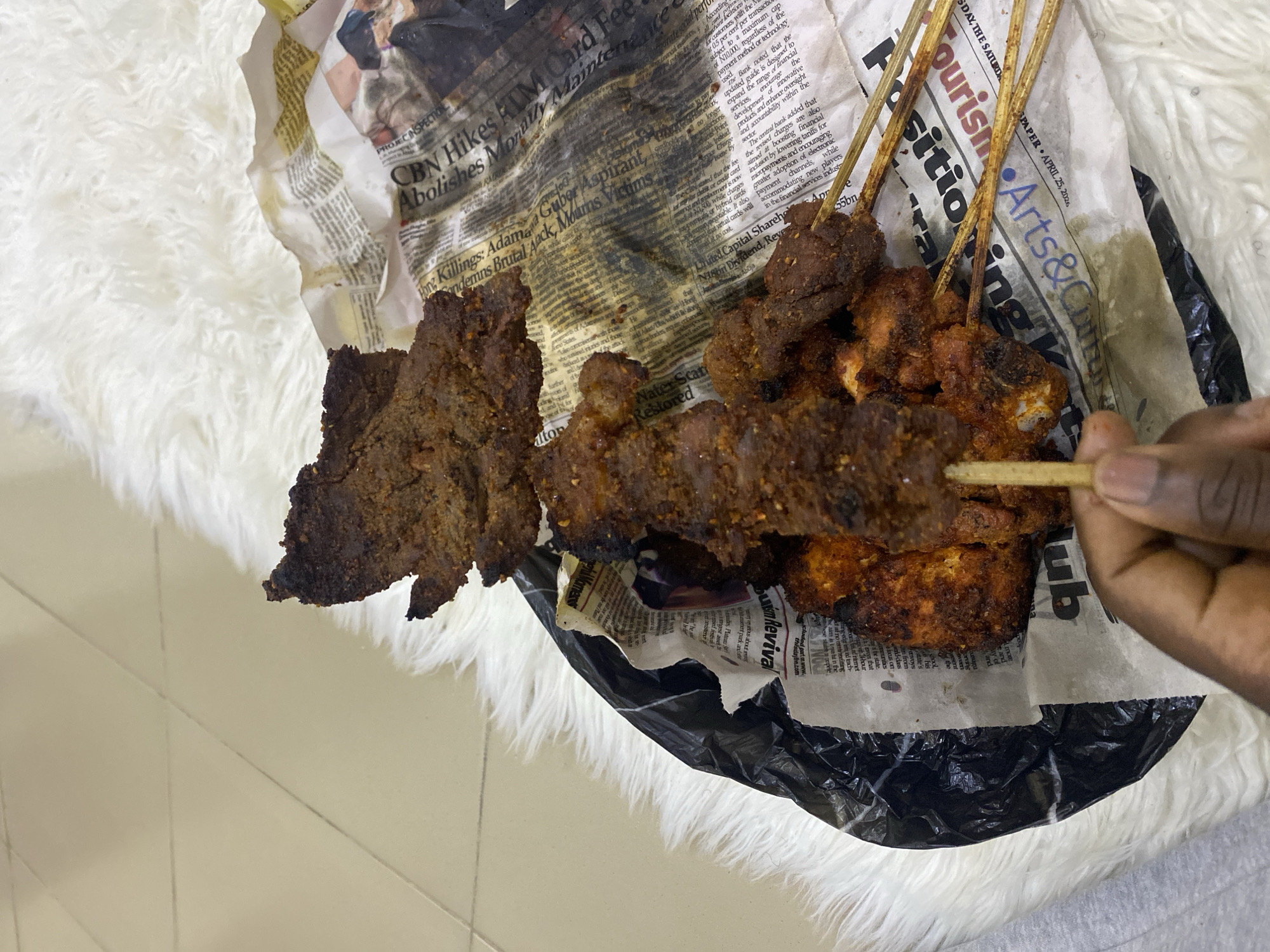 Here's a description of the image:
The scene depicts grilled meat, possibly suya, presented on sheets of newspaper. The newspaper's headlines and articles are partially visible, including mentions of "CBN Hikes ATM Card Fee," "Guber Aspirant Mourns Victims," and "Arts & Culture Club." A hand is holding one of the skewers. The meat and newspaper rest on a white furry surface. The background is a beige tiled floor. There is no direct confirmation of Jos, Nigeria in the image itself; this would require additional information or context.