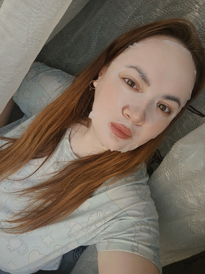 A single female individual is depicted in a medium-shot, from the chest up, reclining in an indoor setting. She has fair skin and long, reddish-brown hair cascading over her left shoulder and chest. Her face is covered with a white sheet face mask, featuring cutouts for her eyes, nose, and mouth. Her eyes are open, brown, and directed towards the viewer. Her lips, visible through the mask, are full and slightly pursed. A small gold-colored hoop earring is visible on her right earlobe. She is wearing a light-colored t-shirt patterned with faint grey outlines of cartoonish shapes. Behind her head is a light blue pillow with a similar subtle pattern. To her upper right and lower left, textured, soft fabric surfaces, likely bedding or upholstery, are visible, predominantly in shades of grey and white. The specific location within the city of Chaikovsky, Russia cannot be determined from the visual information provided.