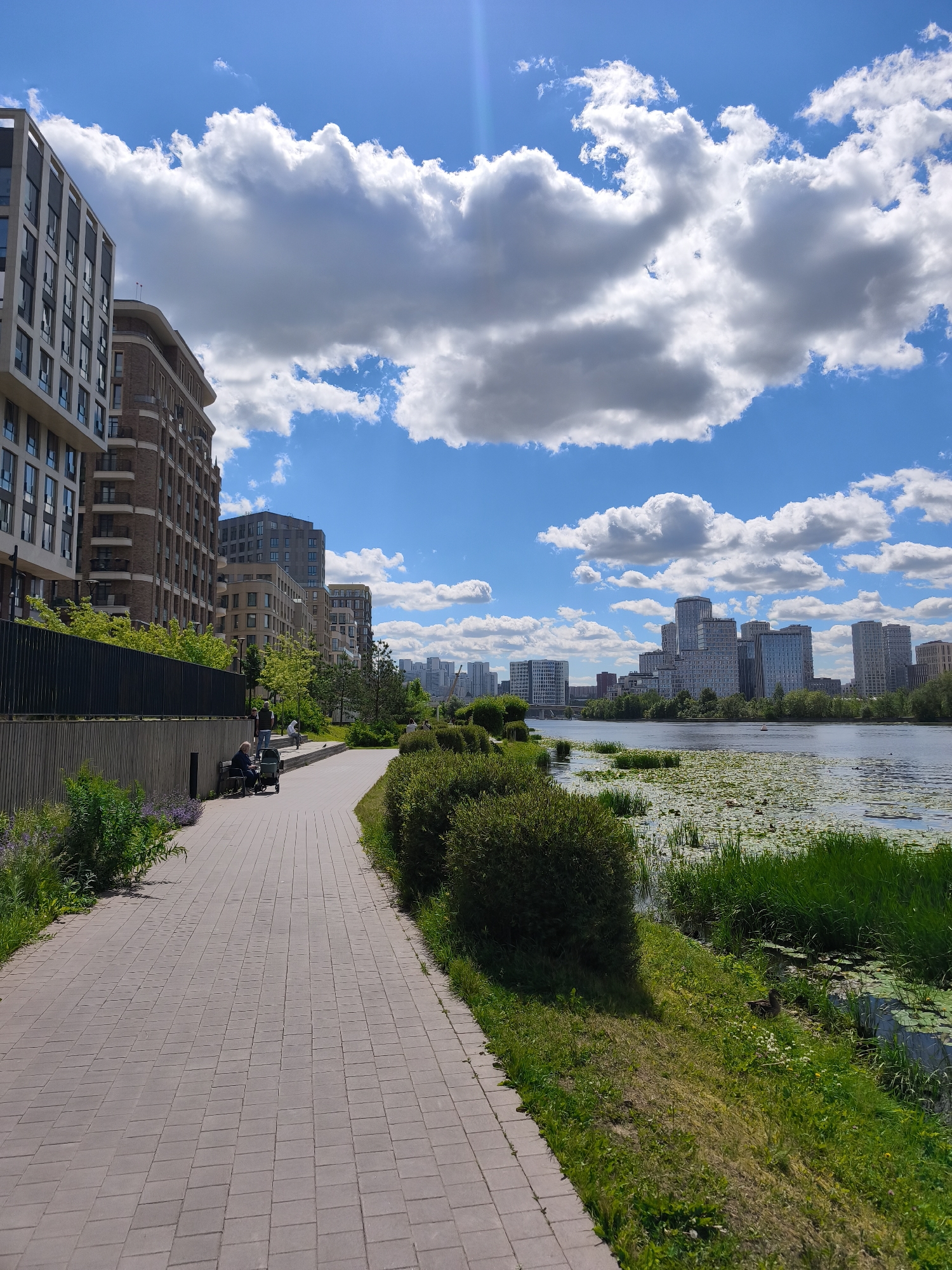 The image depicts a sunny day in Moscow, Russia, likely in a modern urban park area. A paved walkway, constructed from rectangular interlocking stones, curves to the right, bordered by lush green grass and neatly trimmed bushes on the right. On the left, a dark fence separates the walkway from a row of contemporary apartment buildings with multiple stories, featuring modern architecture with large windows and balconies. Further in the background, across a calm body of water covered with lily pads, a skyline of high-rise buildings dominates the horizon. The sky is a vibrant blue, adorned with scattered, fluffy white clouds. In the foreground, on the left side of the walkway, a person sits on a bench next to a stroller, with another person standing nearby. In the middle ground, a duck is visible on the grassy bank near the water's edge. The overall scene conveys a sense of urban tranquility and modern development.