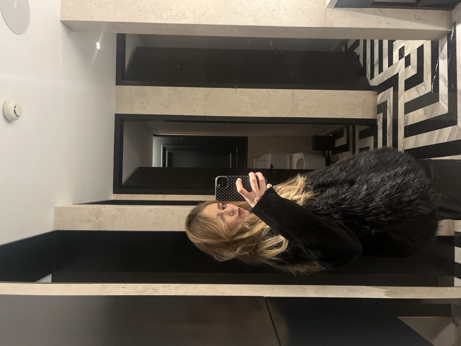 A blonde woman stands in a bathroom taking a selfie in the mirror. She wears a black sweater and a black faux fur jacket. The bathroom features black and white marble floors, black framed mirrors, and white walls. A smoke detector is visible on the upper left wall. The woman holds a black phone case with white polka dots.
