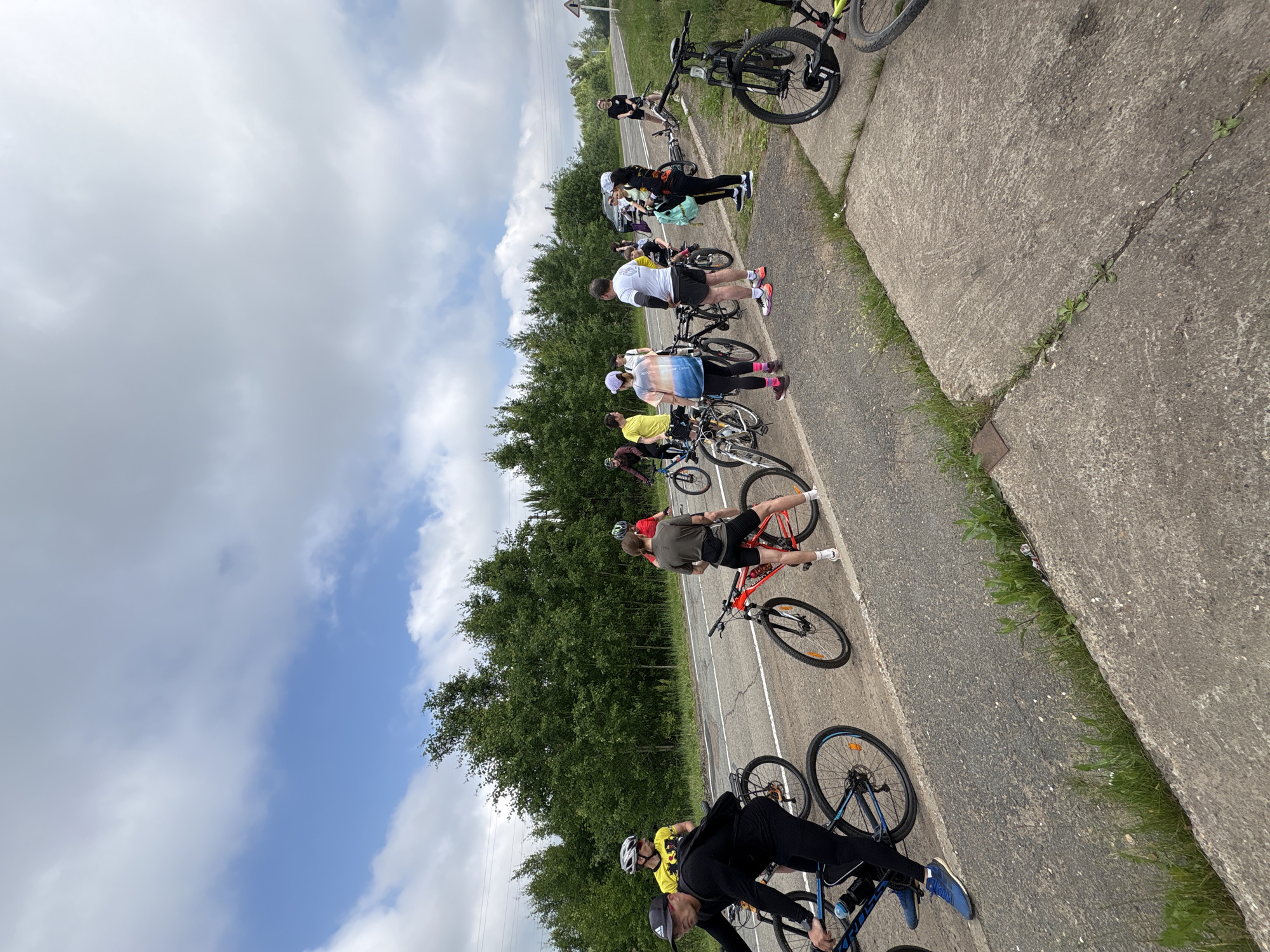 A group of cyclists, numbering approximately twelve, are gathered on the side of a paved road lined with trees. The cyclists, both male and female, are dressed in athletic wear and some are wearing helmets. They are standing near their bicycles, which are parked on the roadside. One cyclist, a male in a black top and black cap, is in the foreground, on the left side of the image, next to his blue bicycle. Another cyclist, a male in a yellow shirt and black shorts, is standing further back, also near his bicycle. A woman in a colorful t-shirt and black leggings is also visible, with her bicycle. The environment appears to be a rural or suburban area, with a clear sky and some clouds. The road has white lane markings. In the background, there is a black car parked on the road. The ground is a mixture of paved road, dirt, and grass. There are also some concrete slabs on the side of the road. The overall scene suggests a break or gathering point for a cycling tour. The location is identified as Kharnavy, Russia.