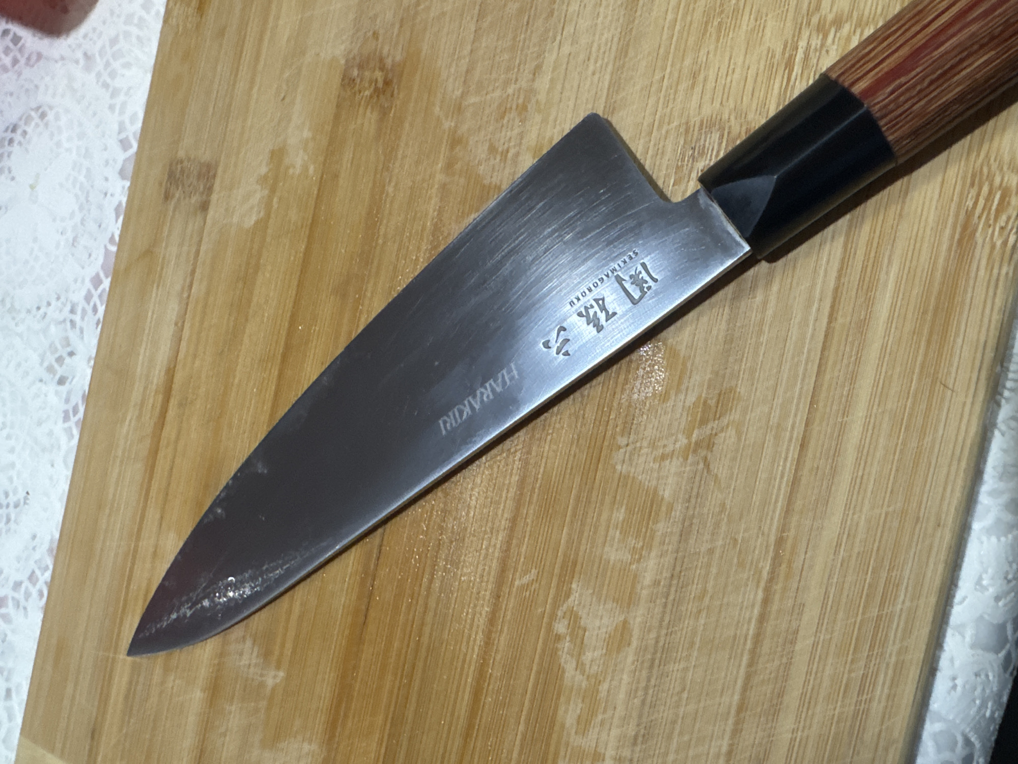 A Japanese-style knife is lying on a wooden cutting board. The knife has a silver blade with Japanese characters and the word "HARAKIRI" printed on it. The handle is made of wood with a black bolster. The cutting board has visible scratches and indentations from previous use. Part of a white lace tablecloth is visible on the left side of the board and another on the right side. The location is not discernible from the image.