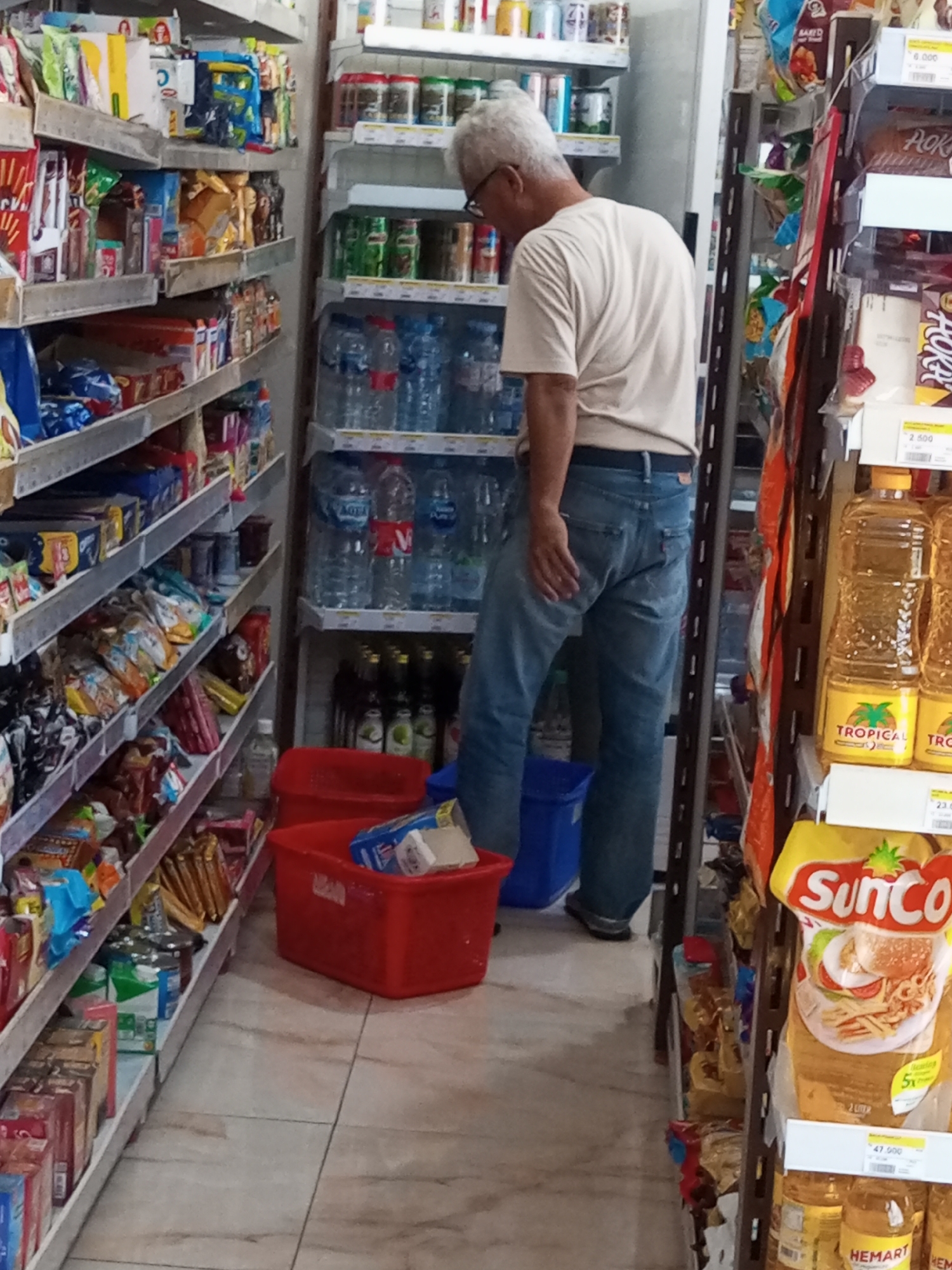 The image shows a man in a light-colored t-shirt and blue jeans standing in a convenience store, looking down at a red bin. He has grey hair and is wearing glasses. The store has shelves stocked with various food items, snacks, and drinks. In the foreground, there are two red bins. One is filled with a white box and other items. To the left of the bins, a shelf is laden with packaged snacks. To the right of the man, there are shelves of bottled water and other beverages. Further to the right, there are bottles of cooking oil and a package of SunCo brand food. The floor is tiled with a marble pattern. The lighting appears to be fluorescent, typical of a retail environment. The image captures a moment of a person interacting with the store's merchandise.